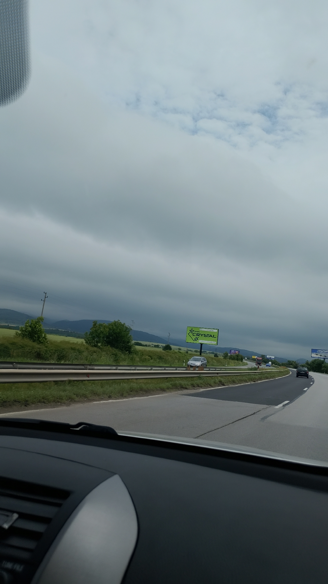 The image captures a view from inside a vehicle on a highway. The sky is overcast with heavy gray clouds, suggesting an impending storm or inclement weather. On the right side of the highway, there is a green billboard with the word "CRYSTAL" prominently displayed in white letters, along with a logo. A silver car is parked on the shoulder near the billboard. The highway itself is paved with asphalt, and a metal guardrail runs along the side. Beyond the guardrail, there is a grassy field and some trees. In the distance, rolling hills or mountains are visible under the cloudy sky. The interior of the car, specifically the dashboard and windshield, are partially visible in the foreground, indicating the perspective of the shot. Several other cars are seen driving on the highway in the distance.