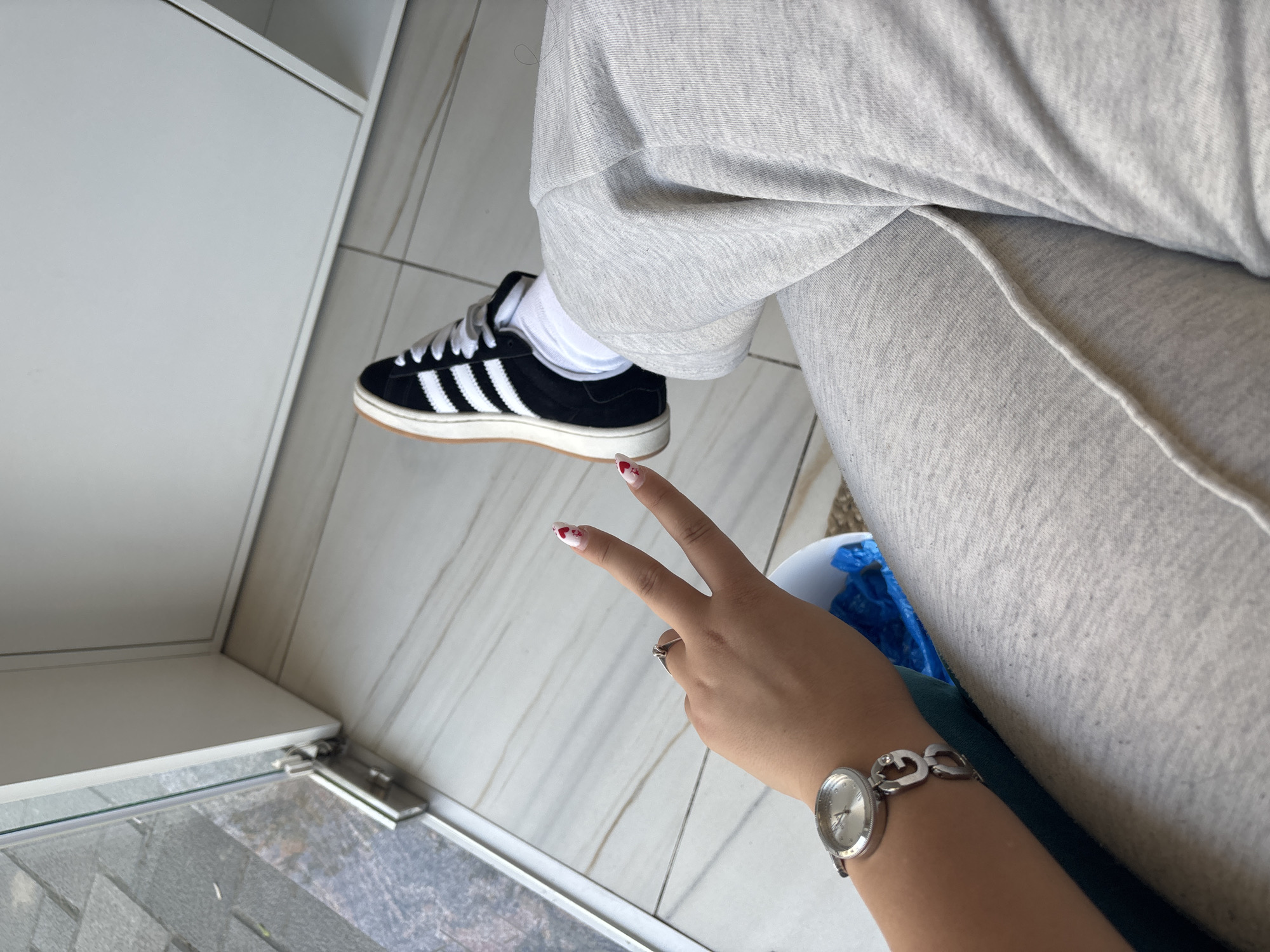 A person is sitting indoors, wearing light grey sweatpants and black Adidas sneakers with white stripes and gum soles, positioned on a white and grey tiled floor next to a white cabinet. The person's left hand, adorned with a silver watch, displays the peace sign. The fingernails are painted white with red hearts. The location is not discernible as being in the city of Plovdiv, Bulgaria, based on the visual information provided.