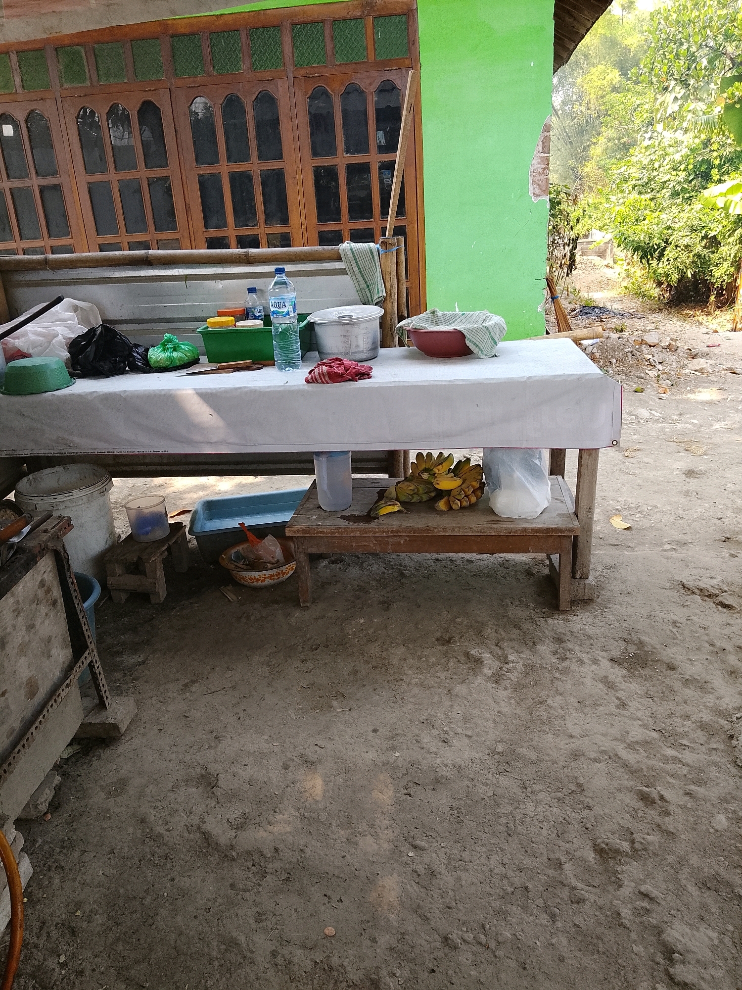This is a photograph taken in Banaran, Indonesia, depicting an outdoor scene in front of a simple building with a green painted wall and large wooden-framed windows. In the foreground, a white tablecloth covers a rustic wooden table. On the table, there are various items, including a bottle of AQUA water, a green plastic container, a bowl with a striped cloth inside, and a bunch of bananas. Underneath the table, on a lower shelf, more bananas are visible, along with a white plastic bag. To the left of the table, there is a large white bucket. The ground is dirt and gravel. The overall setting appears to be a humble outdoor marketplace or roadside stall.