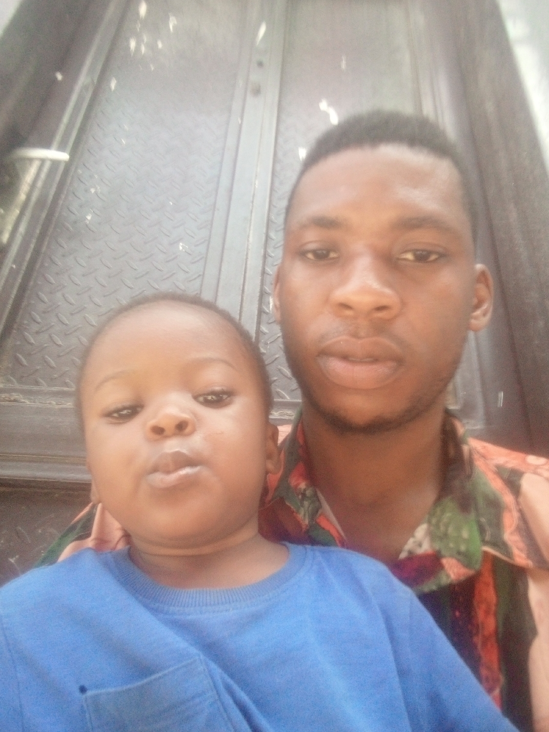 An eye-level close-up shows an adult male and a young child posing for a photograph, likely a selfie. The adult male, with dark skin and short dark hair, is positioned behind and slightly to the right of the child. He is wearing a button-up shirt with a vibrant, abstract pattern featuring shades of orange, green, and black. His expression is neutral as he looks directly at the camera.
In the foreground, a young child with dark skin and short hair wears a plain blue long-sleeved shirt with a small pocket on the left side of the chest. The child's lips are puckered into a slight pout, also looking towards the camera. The child's face fills the lower left and central part of the frame.
The background consists of a metallic door or wall with a raised diamond plate texture, suggesting an industrial or utilitarian setting. A white handle or latch is visible on the far left side of the background. The lighting appears soft and even, typical of indoor or shaded outdoor conditions during the daytime. There is no visible text in the image.