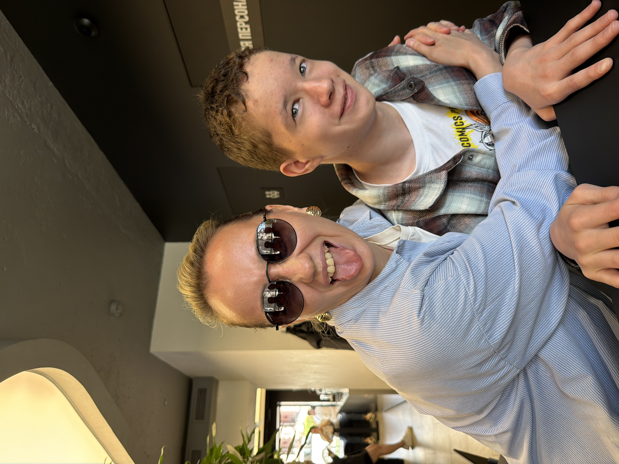 Two people are posing for a photo. A woman with blonde hair and sunglasses is in the foreground, sticking her tongue out. Behind her is a younger man with curly brown hair, wearing a plaid shirt over a white t-shirt. They are indoors, seated at a dark table. The reflections in the woman's sunglasses show other people inside the location. Through a doorway behind her, there is a glimpse of a street scene with greenery. On the table, there is writing in Russian "для персонала" indicating that this might be a staff area. It's not possible to ascertain the location in Izhevsk, Russia from the image.