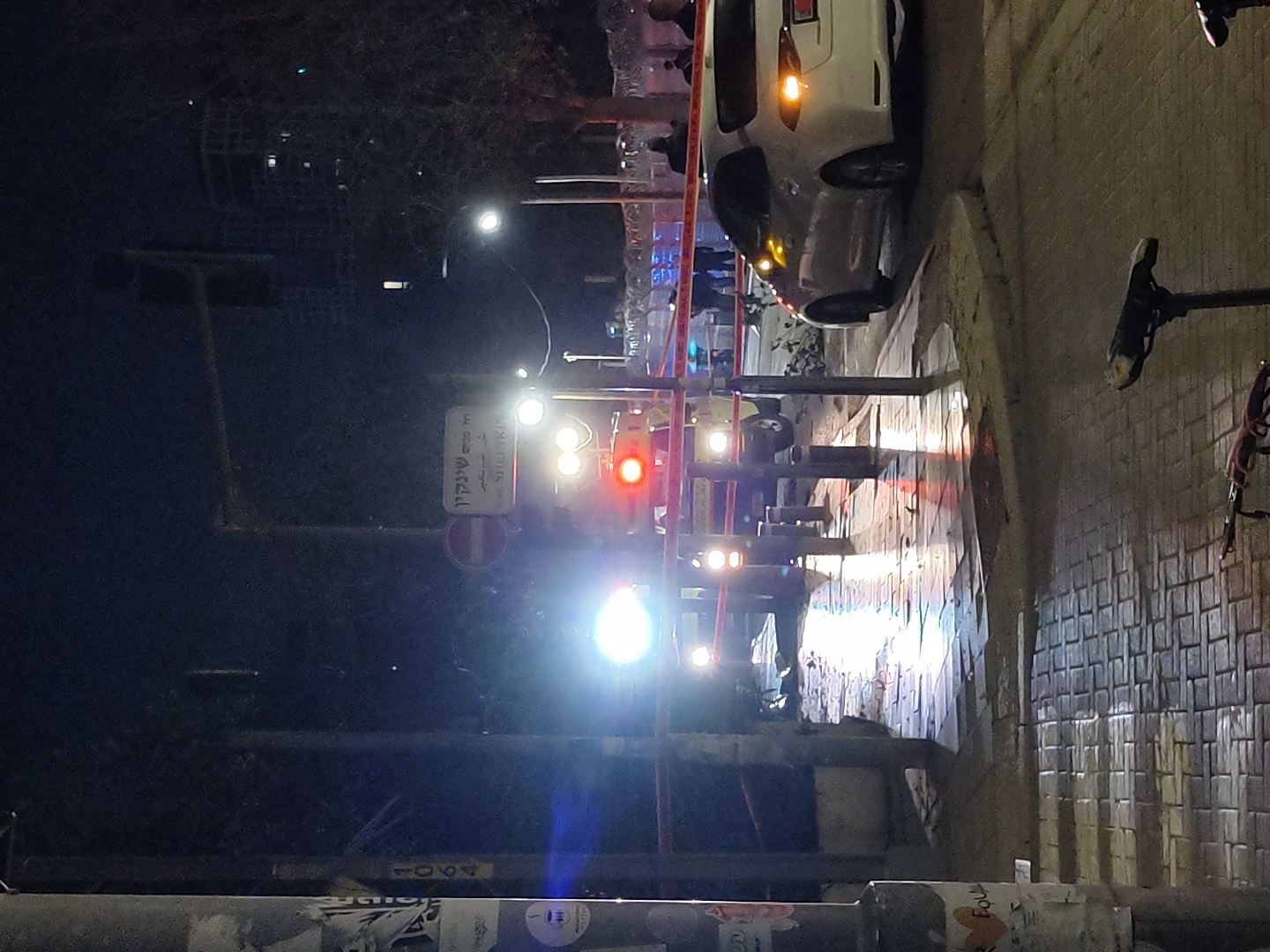 The image depicts a nighttime incident scene on or near Sheinkin Street, at its intersection with Mazini Shlomtzion Street, in Tel-Aviv, Israel.
In the midground, a yellow emergency vehicle, possibly an ambulance or Magen David Adom vehicle, with its bright headlights and red flashing emergency lights activated, is positioned on the wet street. Behind it, additional emergency lights and vehicles are partially visible. A white Toyota sedan is parked on the right side of the street, facing away from the viewer, with its rear lights illuminated.
Red and white striped barrier tape is stretched horizontally across the scene, separating the foreground from the area of the emergency vehicles. Several indistinct figures, presumably emergency personnel, are visible beyond the tape near the emergency vehicles.
The foreground consists of a wet, reflective paved sidewalk and street, reflecting the intense artificial lights from the emergency vehicles and streetlights. A dark utility pole with various stickers, including "1064", stands prominently on the far left. The saddle and handlebars of a bicycle are visible in the immediate bottom foreground. Street signs, including one bearing "SHEINKIN" and "רח' מזיני שלומציון" in Hebrew, are visible above the emergency vehicle. The background reveals dark buildings and bare trees under a night sky.
