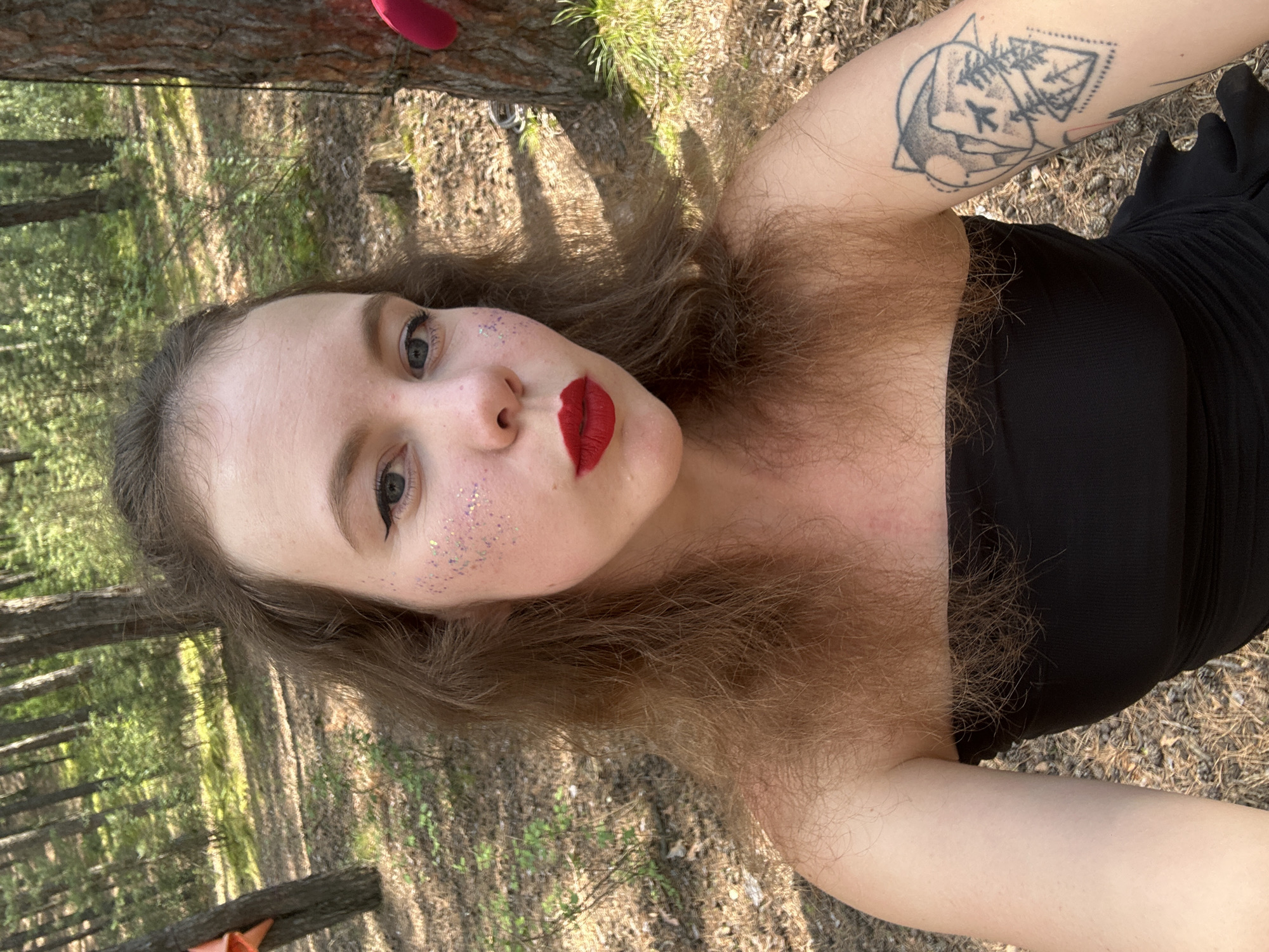 A young Caucasian woman with long, wavy brown hair is taking a selfie in a wooded area. She is wearing a black strapless top and has red lipstick and glitter on her cheeks. She has a tattoo on her left arm. The background shows trees and dappled sunlight on the forest floor. The location is the city of Nikolo-Petrovka, Russia.