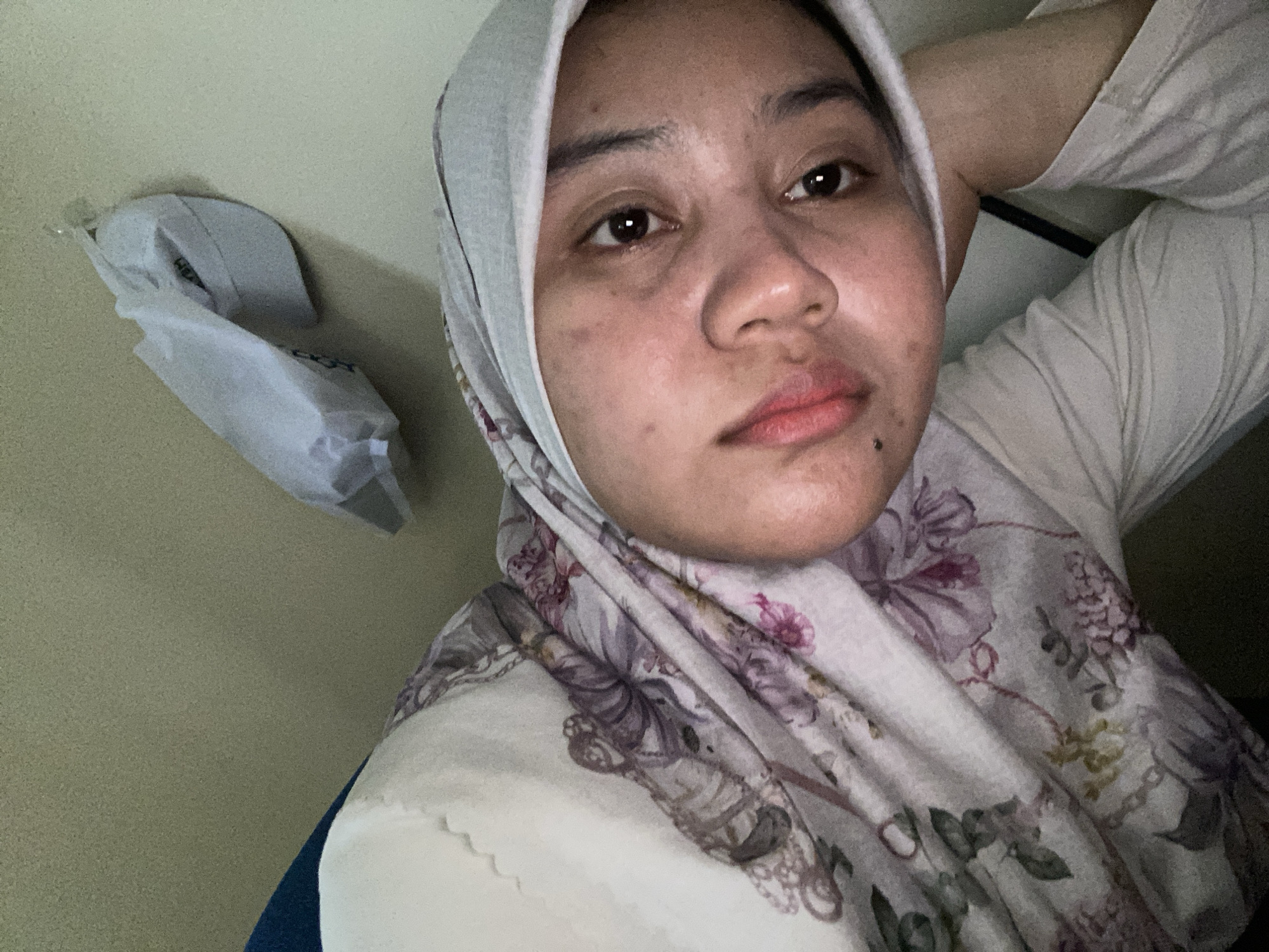 A young woman wearing a hijab and floral print clothing is lying down, resting her head on her arm. She is looking directly at the camera with her eyes open. The background is a plain, light-colored wall with a white object, possibly a folded piece of fabric or a bag, hanging on it. The scene appears to be indoors, likely in a bedroom or living space. The lighting is soft and natural. There are no other people or animals visible in the image. The overall impression is one of a casual and relaxed moment. The location is not discernible from the visual information provided.