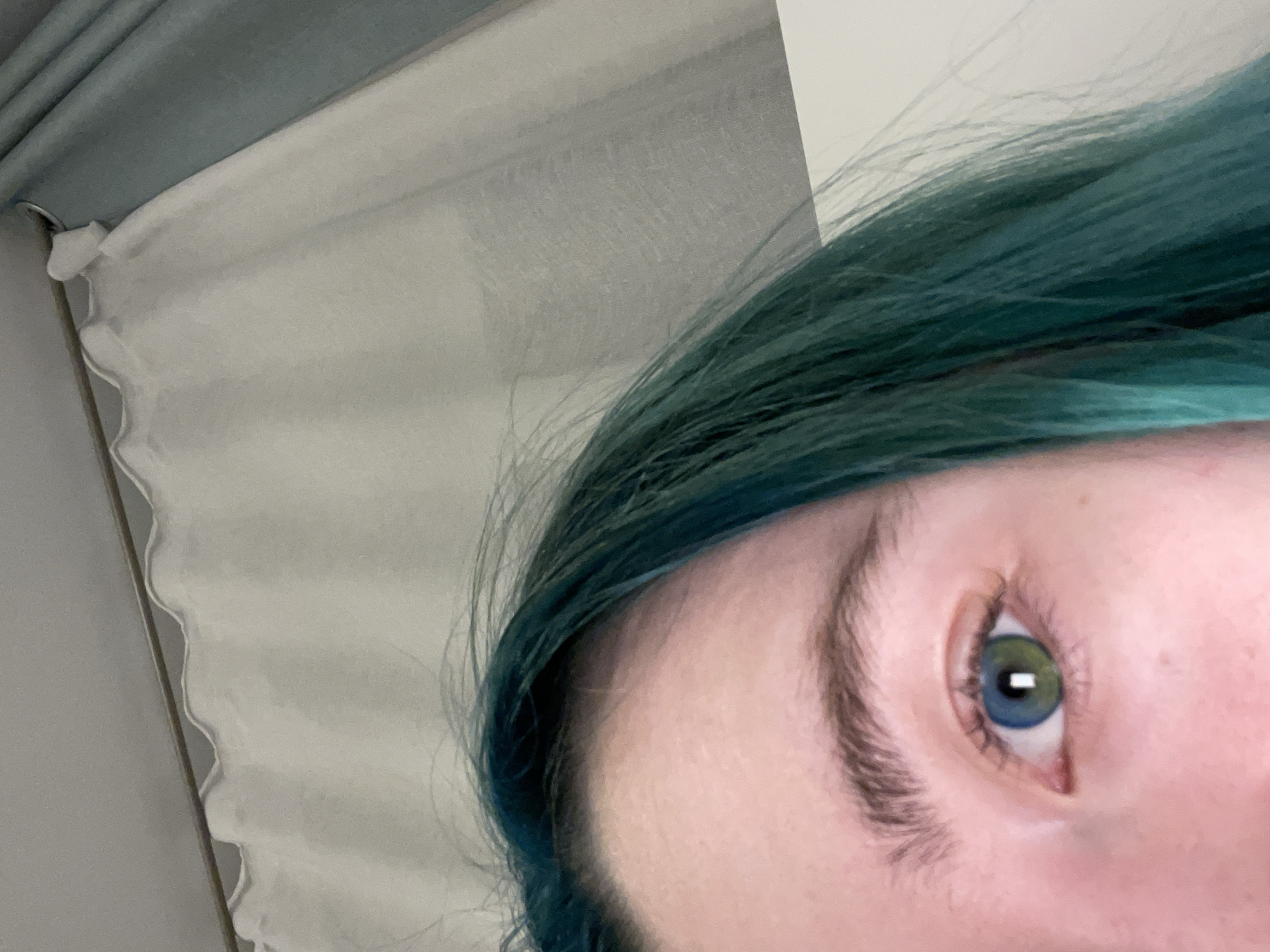 The image shows a close-up of a person's eye and teal-colored hair, with white and grey curtains in the background. The eye appears to have heterochromia, with the outer iris being blue and the inner iris being green. The person's skin is light-colored and appears to have a slight pinkish hue. The hair is long and a vibrant teal color, with some strands falling across the forehead and cheek. The background consists of textured white and grey curtains, suggesting an indoor setting. The image does not contain any identifiable landmarks or details that would allow for its specific location in Tallinn, Estonia, to be determined.