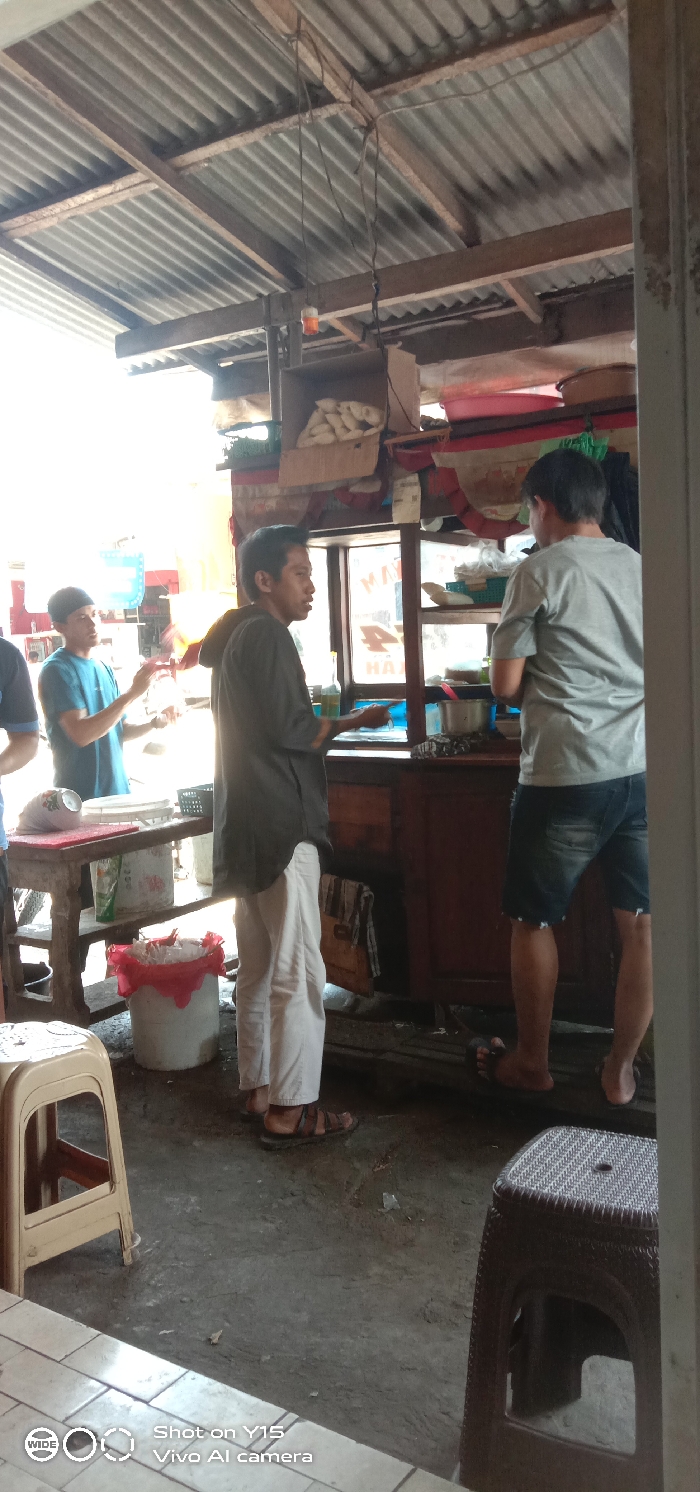 An informal food service area in Maron, Indonesia, is depicted under a corrugated metal roof supported by wooden beams. A single bare light bulb with an amber tint hangs from overhead wiring. On the left, a rustic wooden table with a red cutting board holds a black plastic crate and a white bucket with a red plastic bag liner underneath. A male individual in a blue t-shirt and dark head covering stands behind this table, facing right, with his hands positioned over the table surface. Partially visible to the far left is another individual in a dark blue shirt. In the center, a male individual wearing a dark grey long-sleeved shirt, white trousers, and dark sandals stands facing partially right, looking towards a wooden food counter. His right hand is near the counter. Behind this counter, a male individual in a light grey t-shirt and dark denim shorts, barefoot, stands with his back largely to the camera, actively engaged in tasks behind the counter. The counter features glass display sections and overhead shelves holding a cardboard box with white, elongated food items, along with various plastic basins and containers. The ground is an uneven concrete or dirt surface. A light beige plastic stool is in the bottom left foreground, and a dark brown woven plastic stool is in the bottom right foreground.