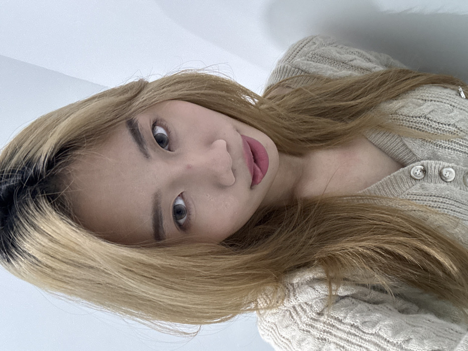 This image depicts a single individual in a chest-up portrait, located within Yuzhong District, China. The subject is female, with an East Asian phenotype and fair complexion. Her hair is long, layered, and dyed a light brown or blonde shade with visible dark roots. She looks directly into the camera, exhibiting wide eyes with apparent light gray contact lenses and defined dark eyebrows. A small red blemish is present on the bridge of her nose. Her lips are adorned with matte pink lipstick, and her tongue is slightly extended and curled upwards. She is dressed in a light-colored, possibly beige or cream, cable-knit V-neck cardigan secured by three visible round, light-colored buttons. The background consists of a plain, uniformly lit white wall, indicating an indoor setting. No other individuals or specific environmental markers are discernible.