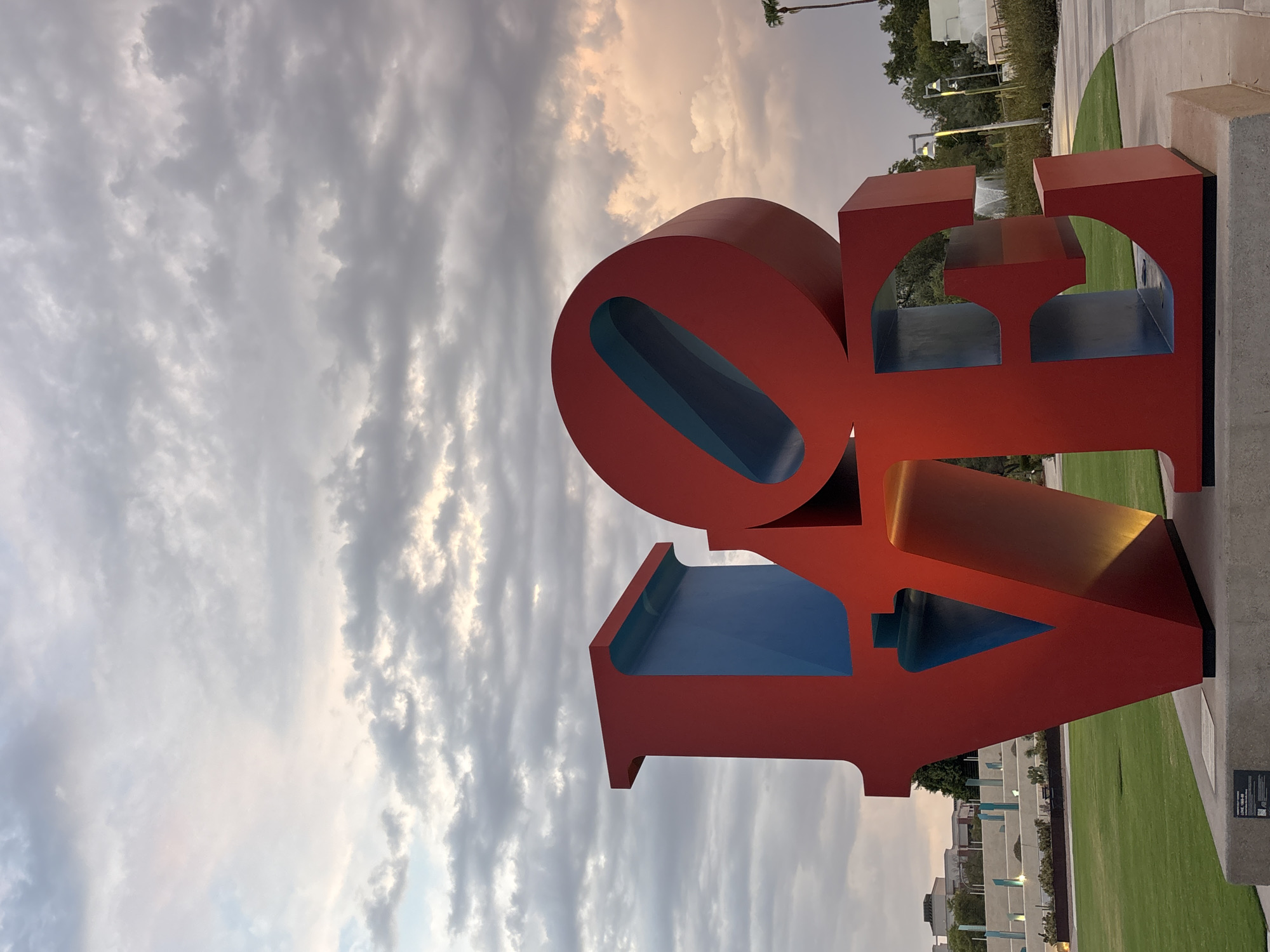 The photograph captures the iconic "LOVE" sculpture in Scottsdale, Arizona, United States of America, at dusk. The large, red, stacked sculpture, illuminated by warm light, is positioned on a concrete base in a landscaped outdoor area. Behind the sculpture, a grassy lawn extends to a paved walkway and a tiered plaza with decorative elements. The sky above is filled with dramatic clouds, transitioning from hues of gray and white to soft pink and orange as the sun sets. Trees and modern buildings are visible in the background, suggesting an urban or park setting. No people are present in the image.