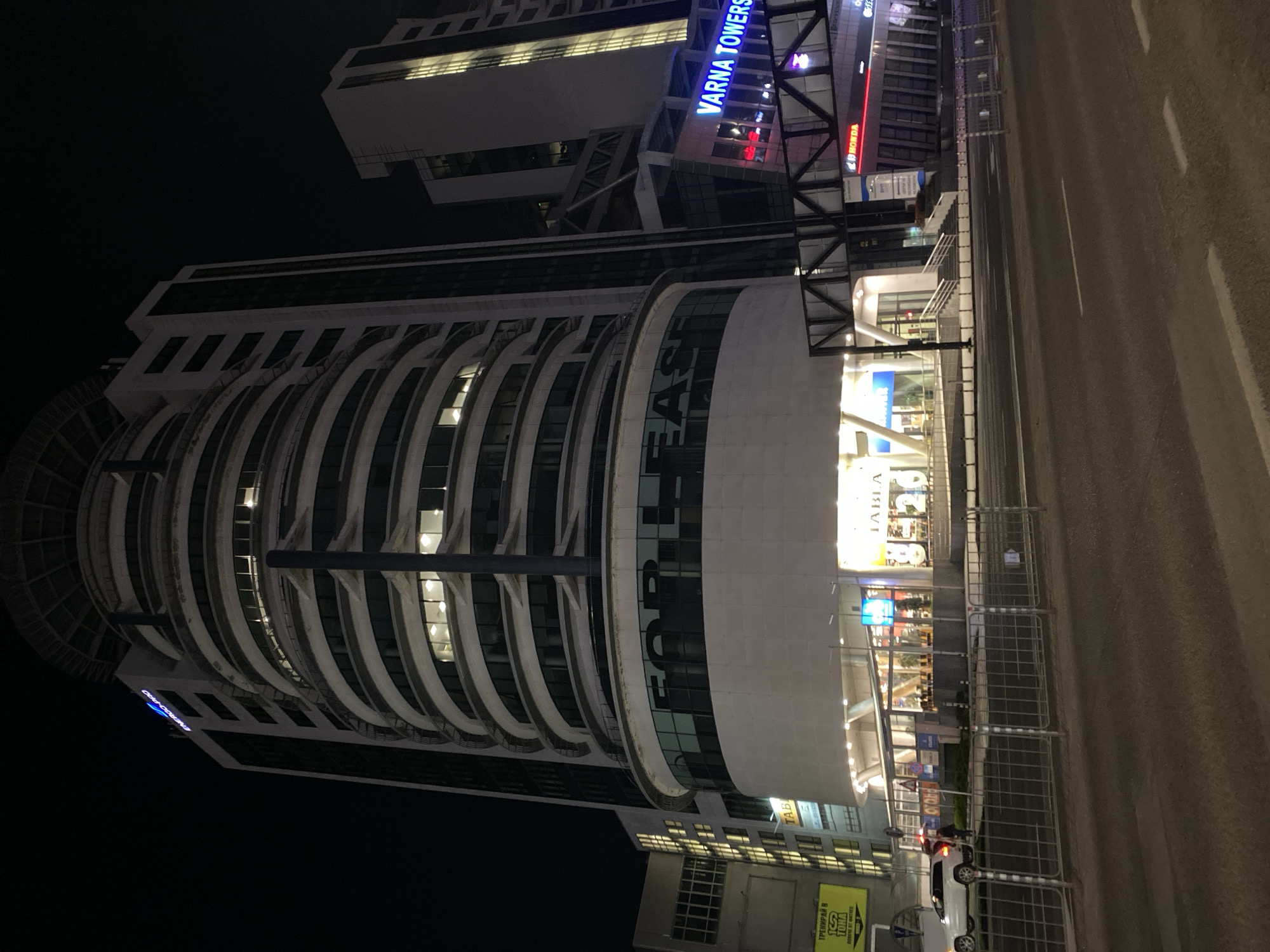 The image depicts a nighttime scene in Varna, Bulgaria, featuring the Varna Towers complex. The illuminated "VARNA TOWERS" signage is visible at the top of one of the buildings. A multi-story building with a curved facade is in the foreground, with the words "FOR LEASE" displayed prominently on its exterior. Streetlights illuminate the adjacent roadway, where pedestrian barriers are set up, and a white car is parked. The scene suggests a commercial district with a mix of retail and office spaces.