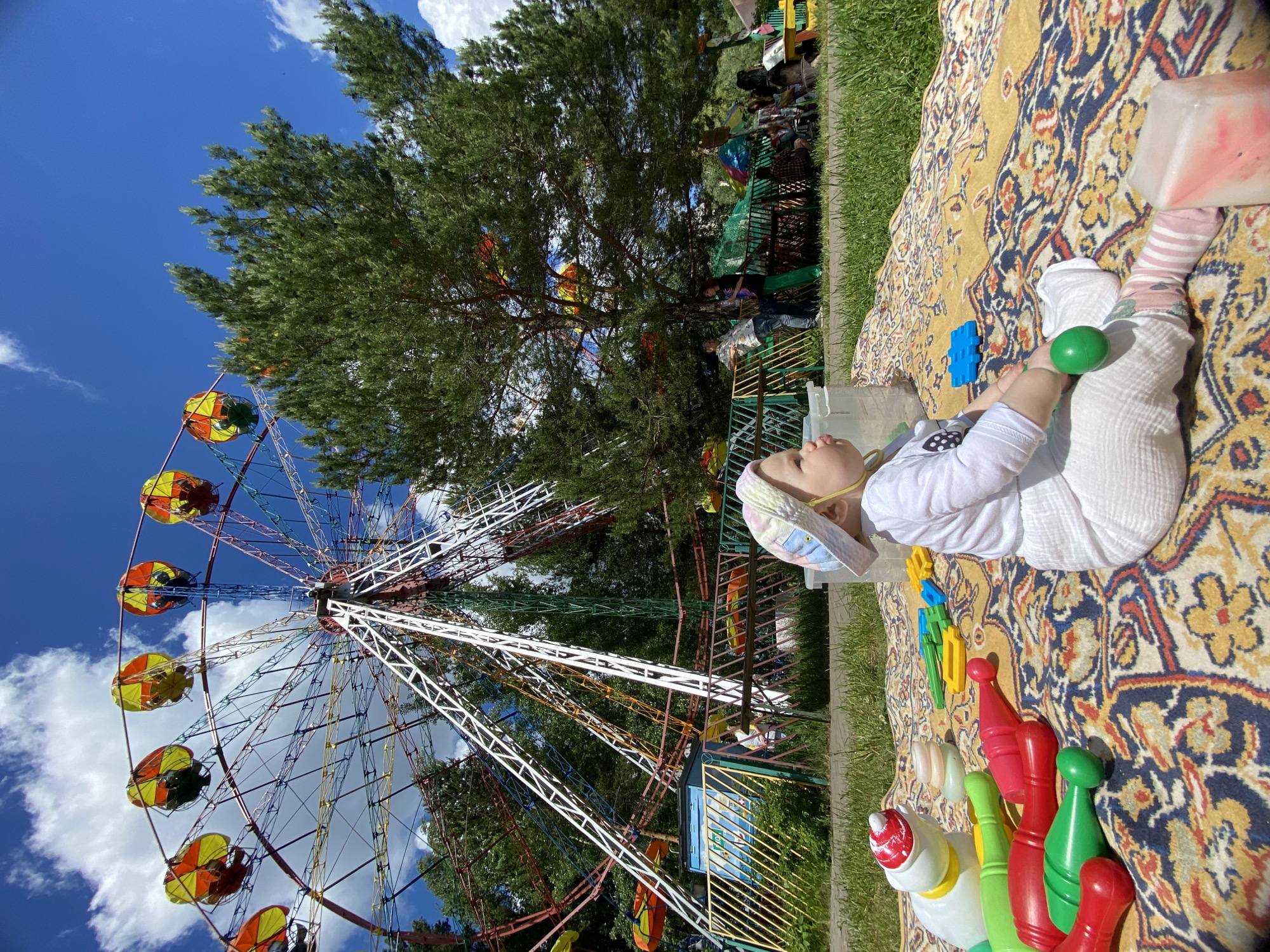 A baby in white pajamas and a patterned hat lies on a patterned rug, playing with toys in a park on a sunny day. The child is surrounded by colorful plastic bowling pins and other toys. Behind the rug is a green grassy area and a fenced-off area with people visible. A large tree and a colorful ferris wheel are visible in the background against a blue sky with scattered clouds. This scene is likely in a park in the city of Chaikovsky, Russia, based on the image data.