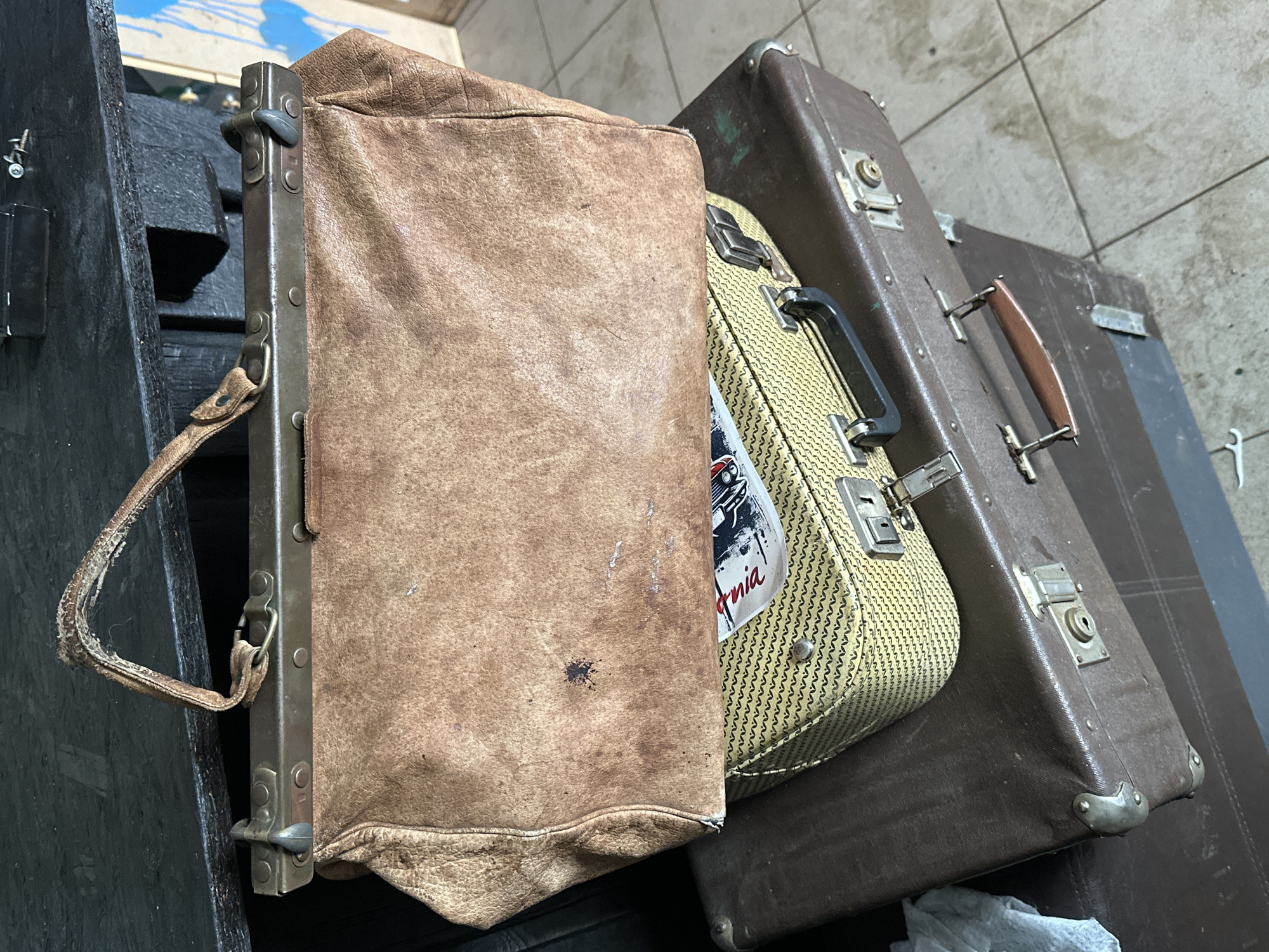 The image shows a close-up of several pieces of luggage. A large, worn brown leather bag with a metal frame is positioned on the left side of the frame. To the right, a smaller, patterned suitcase sits partially obscured by the leather bag, and a larger, brown suitcase is in the background. The luggage is placed on a dark-colored surface, possibly a crate or platform. The floor is tiled with light-colored square tiles. There is no indication of the location being Andreyevka, Russia. The image contains no people or actions.