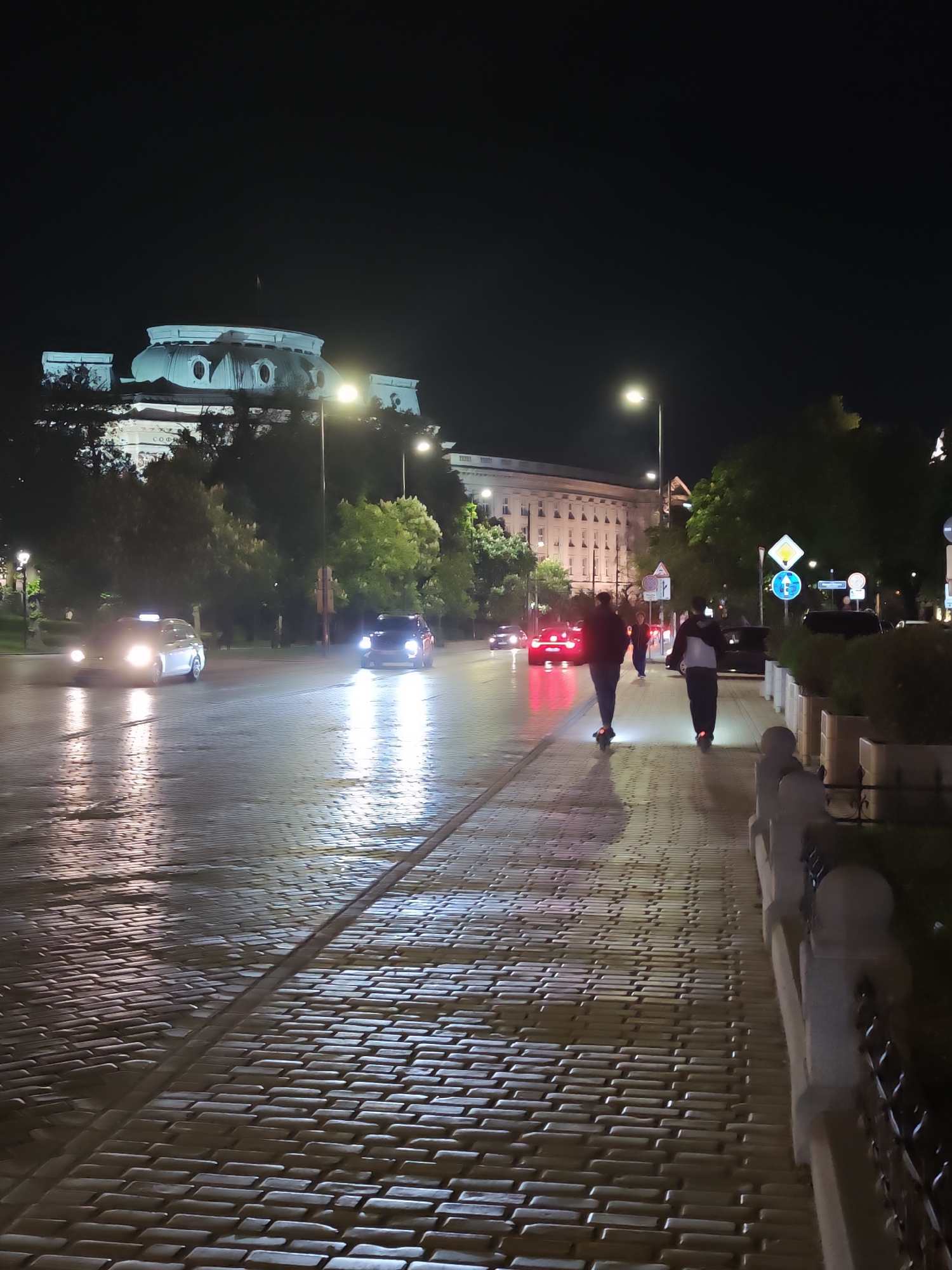 The image depicts a night scene in Sofia, Bulgaria, recognizable by the architecture of the parliament building in the background. The location appears to be a major avenue or street. The cobblestone street and sidewalk are wet, reflecting light from streetlights and passing cars. Several vehicles, including cars and a taxi, are visible on the road. Two individuals are riding scooters on the sidewalk. The scene suggests a rainy or recently-rained condition.