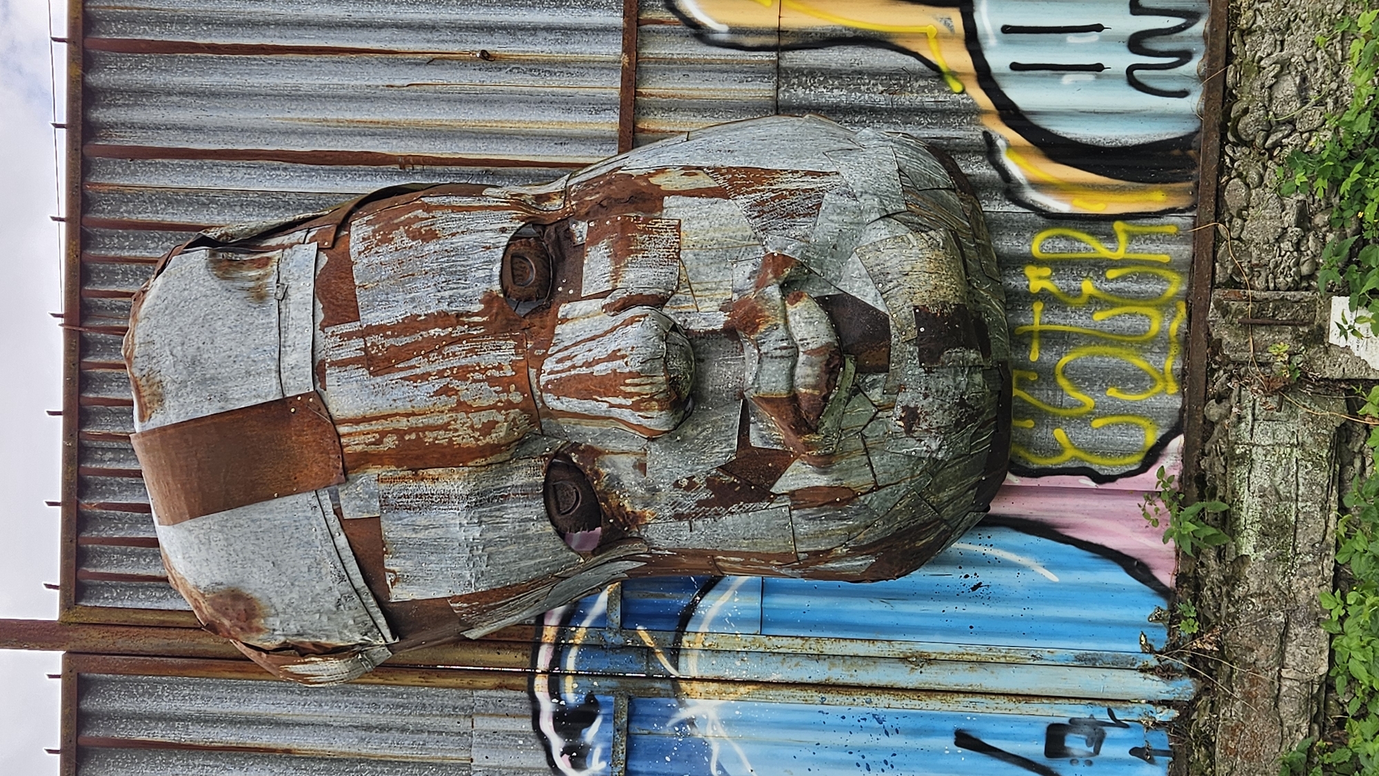 The image shows a large, rusted metal sculpture of a baby's head affixed to a corrugated metal wall. The sculpture is made of various pieces of metal, some of which are heavily rusted, giving it a textured and weathered appearance. The face features deep-set eyes, a prominent nose, and full lips. Behind and to the sides of the sculpture, the corrugated metal wall is painted with graffiti. To the left, there are blue and white wave-like designs with black spray paint accents. To the right, there are yellow letters forming the word "ESOTER" and further to the right, a simplified cartoon face with black lines. The base of the wall rests on a rough stone foundation, overgrown with green vegetation. This artwork is located at the intersection of Fintická and Sekčovská streets in Prešov, Slovakia.