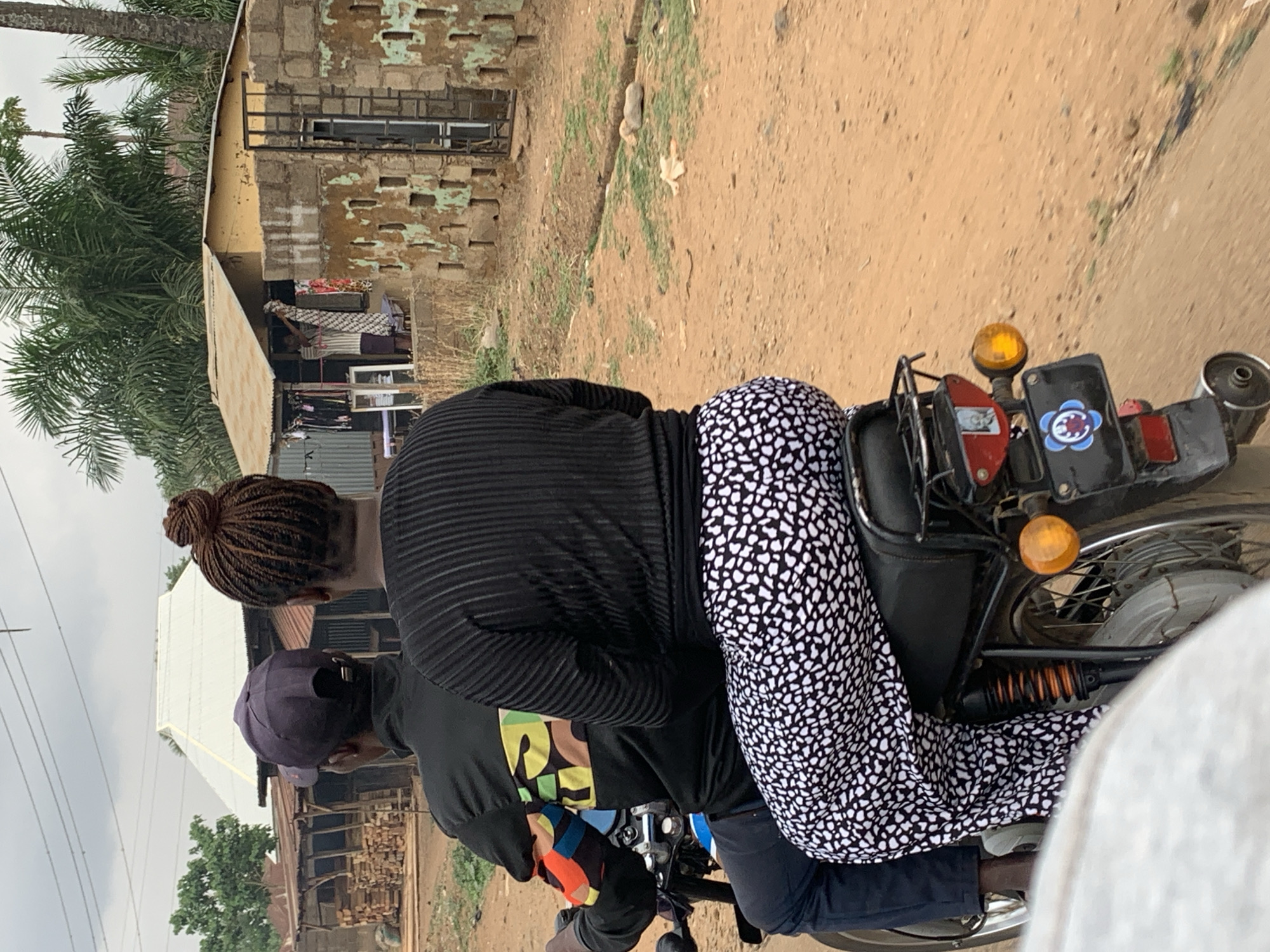 Two people are riding a motorcycle on a dirt road in Maigizo, Nigeria. The person in the back has black-and-white patterned pants and a black shirt, while the driver is wearing a black shirt, dark pants, and a baseball cap. The motorcycle is black with yellow lights and a license plate with a flower sticker. Buildings and trees are visible in the background. The road is made of reddish-brown dirt with some patches of grass.