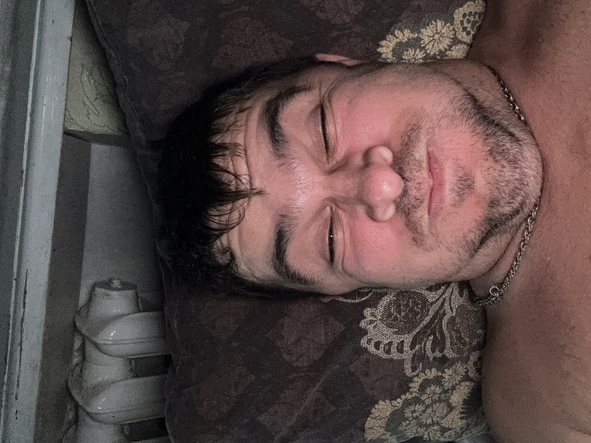 A selfie of a man lying in bed. He is looking at the camera with his eyes squinted and his cheeks puffed out. He is wearing a silver chain around his neck. The man is lying on a patterned pillow, and a radiator can be seen in the background. The image appears to have been taken in a bedroom.