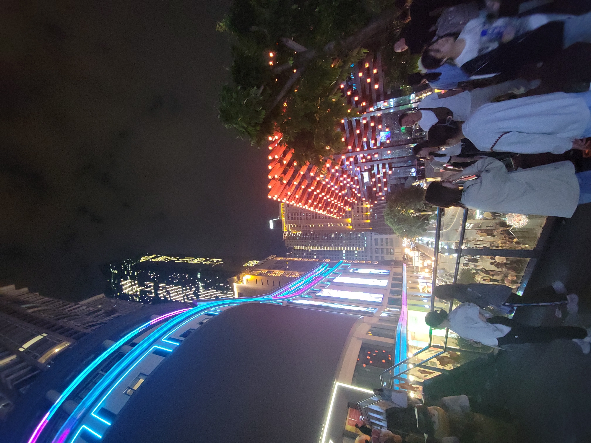 The image captures a vibrant urban night scene in Yuzhong District, China, characterized by extensive artificial lighting and modern architecture. The photograph appears to be taken from an elevated perspective, looking down and across a lively promenade or walkway.
In the foreground and midground, numerous people are visible, engaged in leisure activities such as walking, standing, and observing the surroundings. Many are casually dressed, suggesting an evening outing. A prominent feature is a series of red rectangular light fixtures, resembling modern interpretations of lanterns, illuminating a treed area and an overhead structure. Below this, a long, curved architectural element is strikingly lit with bright blue and pink neon strip lights, creating a dynamic and futuristic aesthetic. Glass railings and partitions separate the walkway from other sections of the area.
In the background, a dense urban landscape unfolds, dominated by tall skyscrapers and other modern buildings. Many of these structures are illuminated with internal lights, showcasing numerous windows and contributing to the city's night glow. The sky is dark, indicating nighttime, and the weather appears clear. No specific events beyond general evening leisure are clearly depicted, but the atmosphere suggests a popular commercial or tourist area.
No legible text is visible within the image. The unique blend of traditional-inspired red lighting and contemporary neon accents, along with the high-rise architecture, strongly cues the modern East Asian urban setting, consistent with the provided location context of Yuzhong District, China.