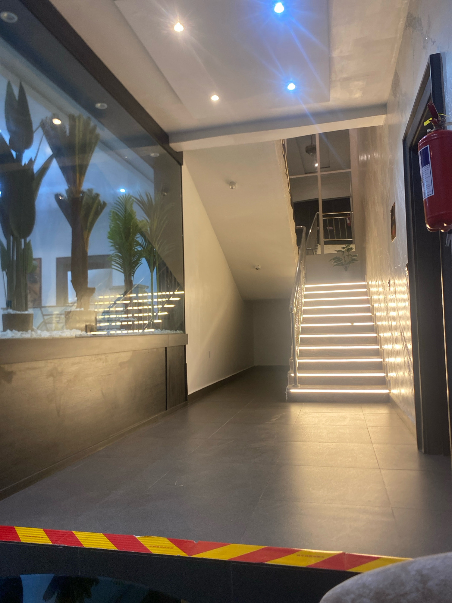 This image depicts a modern indoor staircase in Barden Yana, Nigeria. The staircase is illuminated by warm LED lights embedded in each step, creating a dramatic visual effect. The walls are finished with a textured plaster, and the flooring appears to be dark tiles. To the left, a large glass enclosure houses artificial plants, possibly a decorative element. A red fire extinguisher is mounted on the wall near the top of the staircase, next to a doorframe. A yellow and red striped caution tape is visible in the foreground, suggesting a temporary boundary or hazard warning. The overall impression is one of contemporary design with an emphasis on lighting and texture.