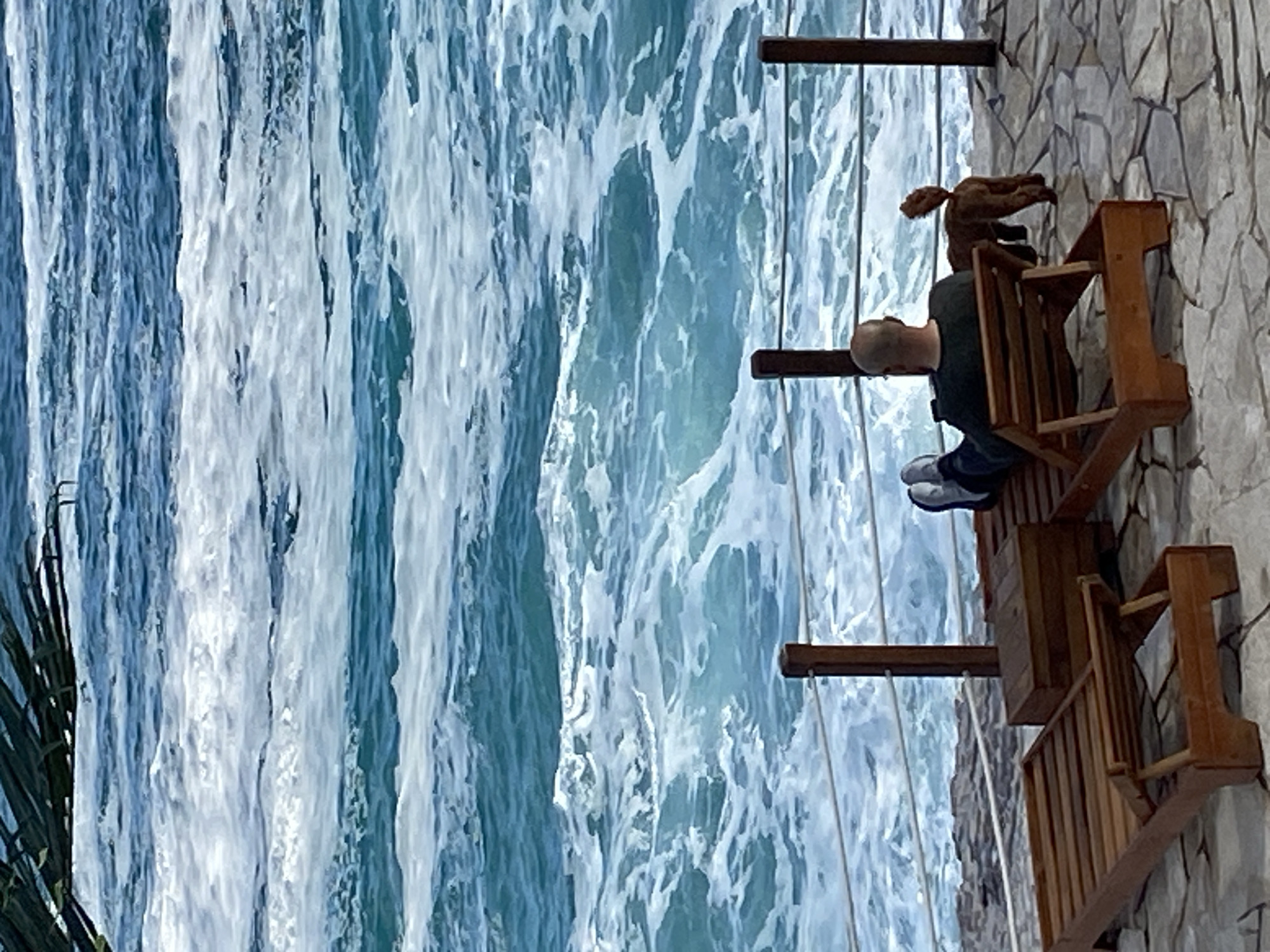 The image depicts a man sitting on a wooden chair on a balcony, overlooking a rushing river or waterfall. The man has a shaved head, is wearing a dark shirt and jeans, and white sneakers. He is looking down at the water. On the back of the chair is a stuffed animal, possibly a dog. The balcony is made of wood and is attached to a stone wall. The background is dominated by the cascading water. The location is indicated to be Prijevor, Montenegro, suggesting the water feature is likely a significant natural attraction in the area. The water appears turbulent, with a mix of blue and white, indicating a rapid flow.
