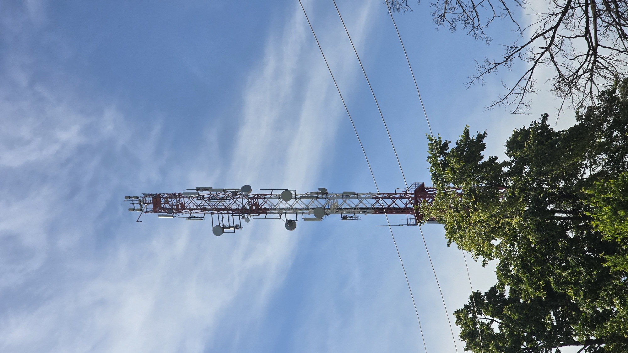 The image shows a tall telecommunications tower against a bright blue sky with wispy clouds. The tower is red and white and is adorned with multiple antennas and dishes. In the foreground, lush green trees partially obscure the base of the tower, and bare branches from another tree are visible to the right. Power lines stretch horizontally across the frame, in front of the tower. The scene is outdoors, with the focus on the technological structure against a natural backdrop. The image does not depict any people or specific urban landmarks to indicate a precise location within the city of Актюбинский, Russia.
