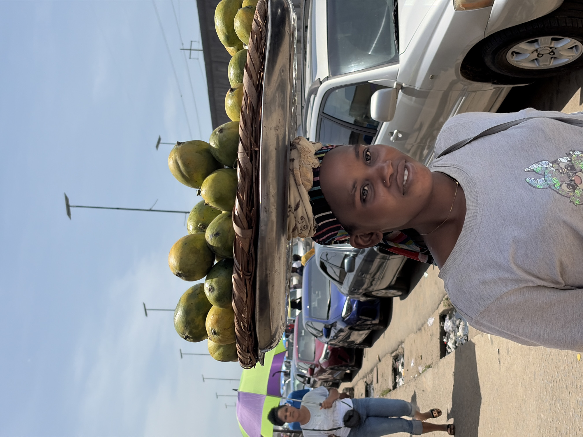 A medium shot captures a woman in Lekki, Nigeria, smiling at the camera while balancing a tray laden with mangoes on her head. The mangoes, stacked high, display a mix of green and yellow hues. The woman is wearing a light gray sweater with a sequined design on the chest. She also has a nose piercing and a thin gold necklace. A striped headwrap covers part of her head. The background reveals a city street with parked cars, including a white SUV. Other people are visible in the distance, including a woman holding a purple and green umbrella. Litter is scattered on the sidewalk. Utility poles are visible against a bright blue sky.
