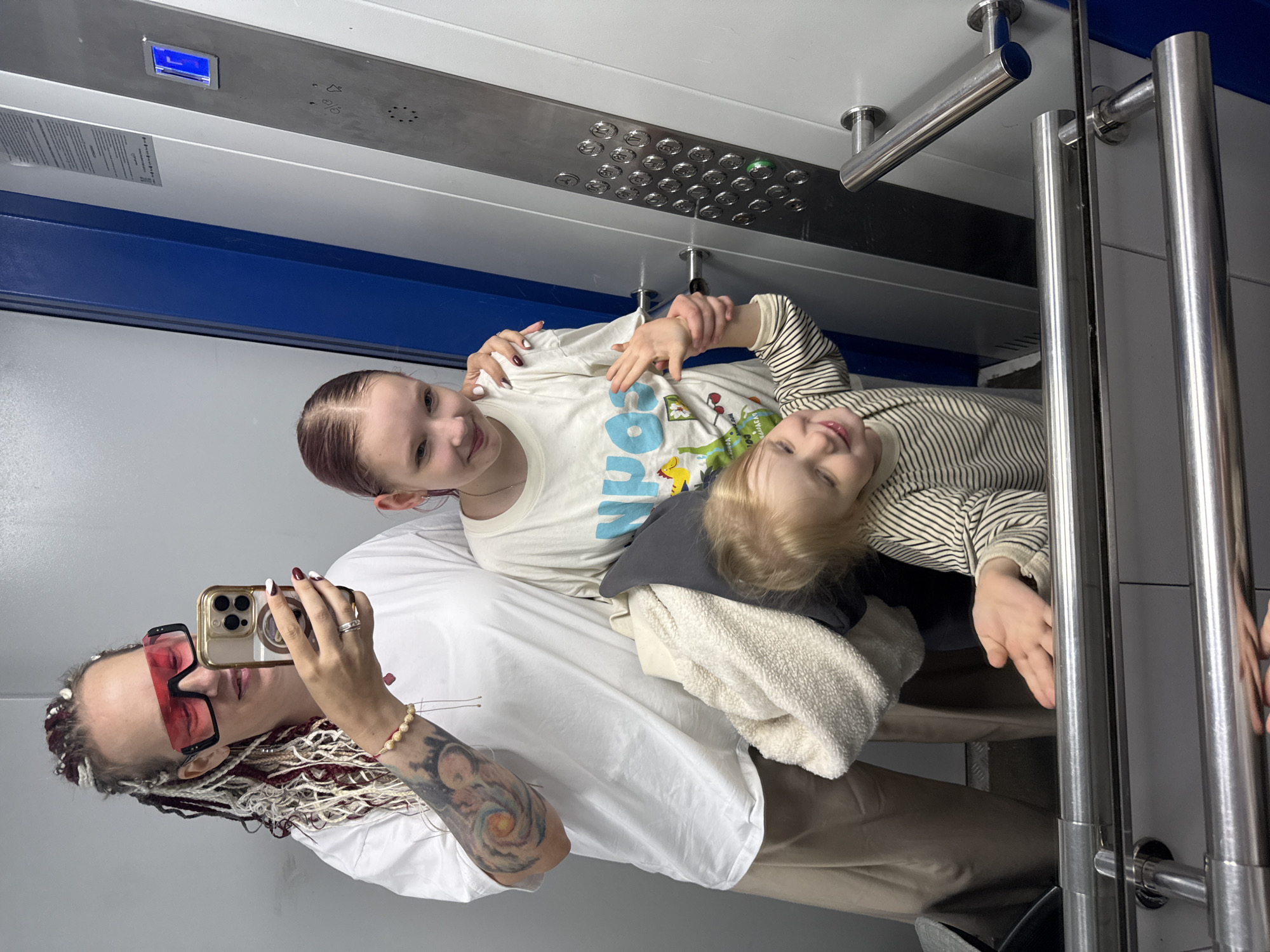 A woman with braided hair and large pink sunglasses takes a selfie in an elevator with two young children. The woman wears a white t-shirt and tan pants, with tattoos visible on her arm. The older child, a girl with reddish-brown hair, stands to the woman's right, looking at the camera. A toddler with blonde hair stands in front of the woman and girl, reaching out with one hand. The elevator has a silver control panel with buttons and a blue illuminated display. The background is a light gray wall.