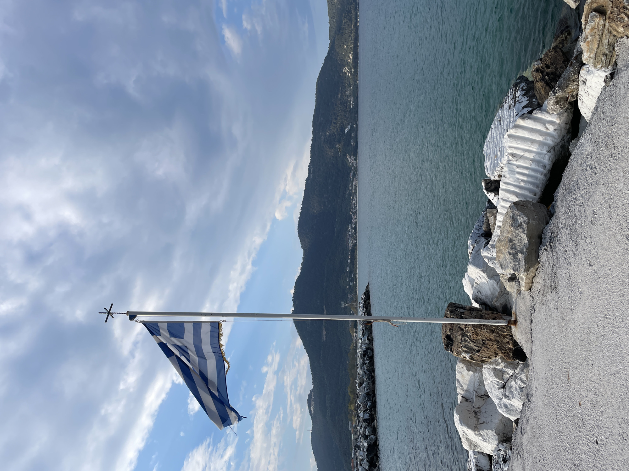 The image is a sideways view of a Greek flag on a flagpole, with a cross at the top, against a backdrop of a cloudy sky, a mountain range, and a body of water. The flagpole is mounted on a rocky barrier, likely a breakwater, made of white and gray stones. The barrier is adjacent to a paved area, possibly a walkway. The location is likely Skala Potamias, Greece, based on the presence of the Greek flag and the coastal landscape. No people or specific actions are visible in the image.