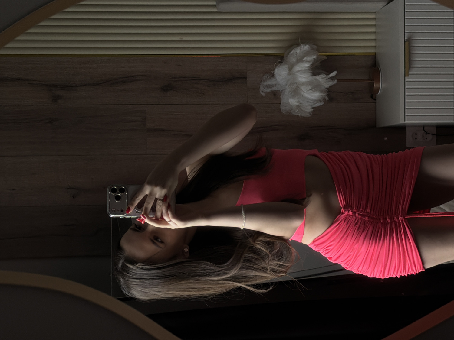 The image captures a young woman taking a mirror selfie in an indoor setting. The scene is viewed as a reflection, with the woman appearing to lie on her back, holding a smartphone (an iPhone with a triple camera setup) to capture her image in a large mirror. She is wearing a bright pink or coral ruched dress. Her long brown hair is spread around her head, and she has red nail polish and a ring on her left hand, along with a bracelet on her right wrist.
The background visible in the reflection shows a wooden floor. To the right of the woman, a light-colored cabinet or dresser with horizontal slatted details is visible, along with an electrical outlet featuring two sockets. A white, feathery decorative object, possibly a lamp, is positioned on the floor near the furniture.
The lighting suggests an indoor setting, possibly with artificial light, as the woman's reflection is brightly illuminated, creating a strong contrast with the darker surrounding room. Her expression is neutral, focused on taking the photograph. There is no visible text in the image.