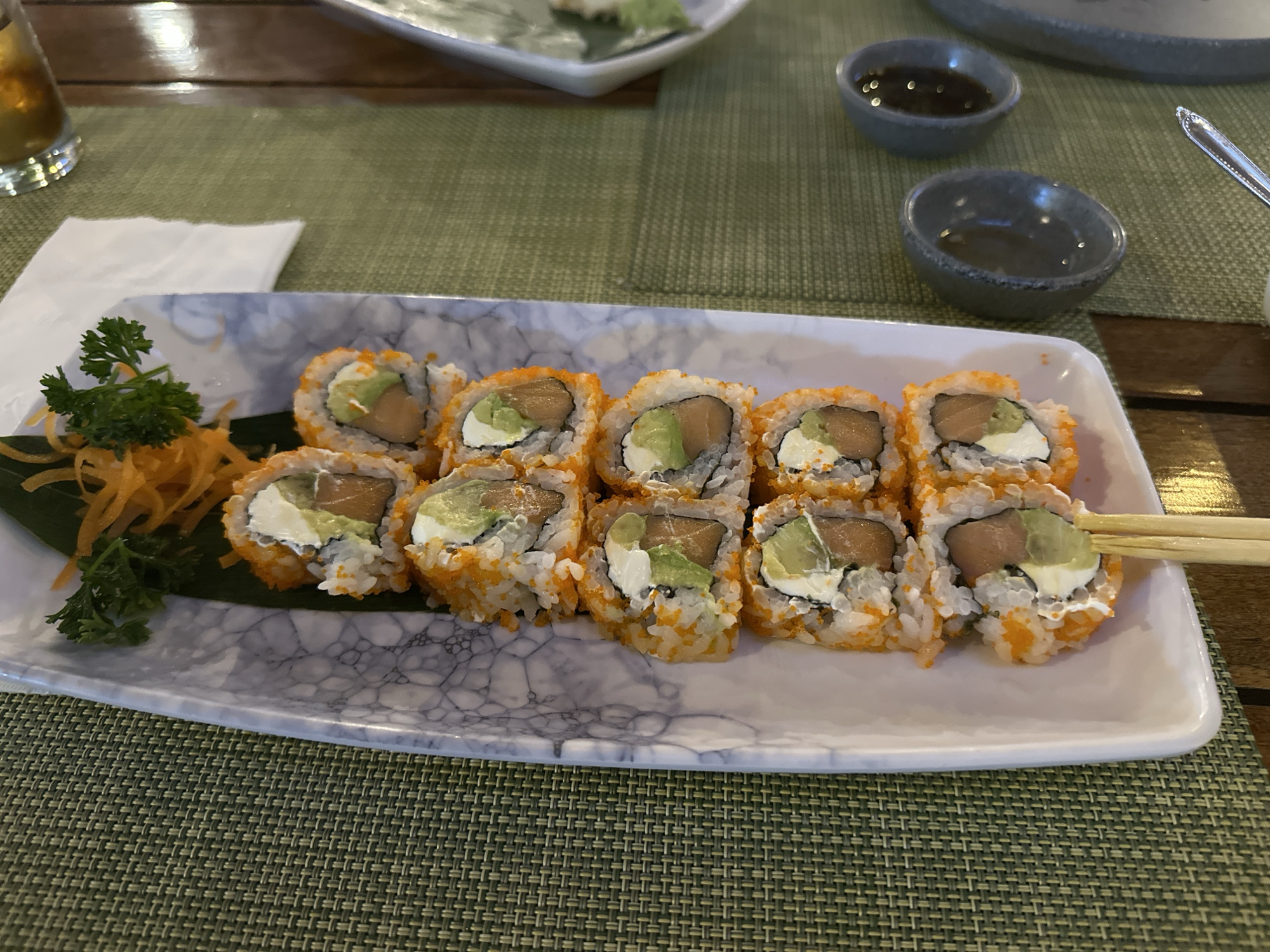 A plate of sushi rolls is on a table. The sushi rolls appear to be California rolls with salmon and avocado. The plate has a white marble pattern. The table has a green woven placemat. Two small bowls with sauce are visible. A glass of beer, a plate with food, and a fork are in the background. The image was taken indoors. The location is not verifiable as San Bernardino Tlaxcalancingo, Mexico, without additional evidence.