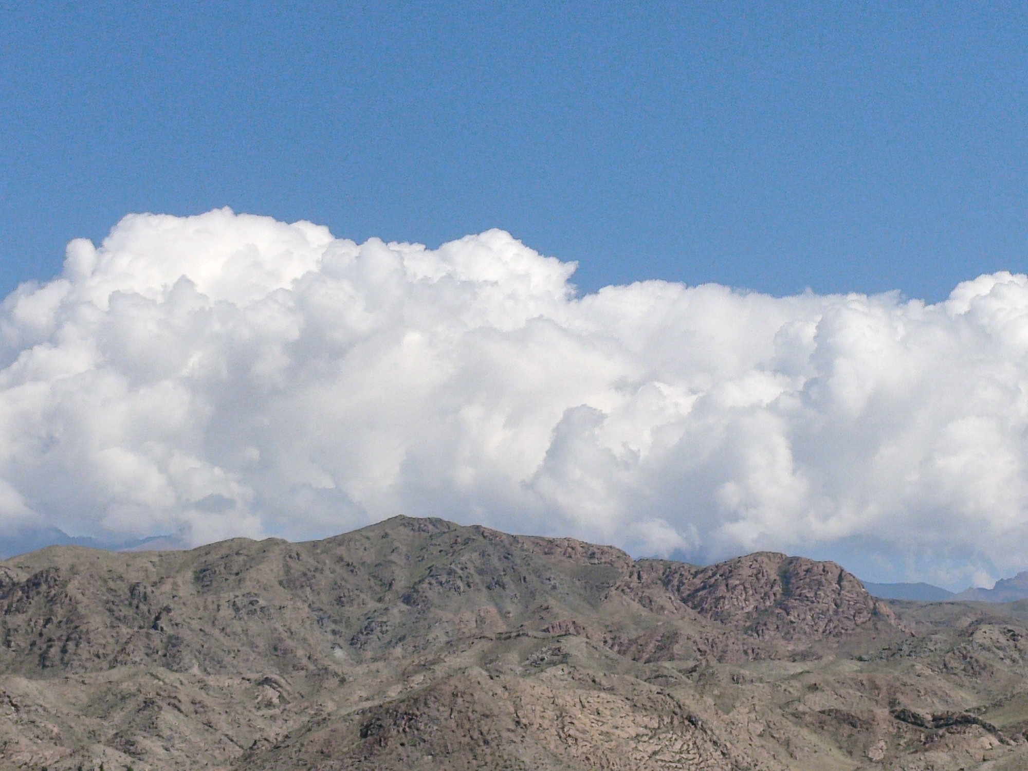 A landscape photograph displays a mountain range under a bright blue sky filled with dense, fluffy white clouds. The mountains are rugged and rocky, with varying shades of brown and green. The clouds form a dramatic line across the upper portion of the image, partially obscuring the sky above the peaks. The scene appears to be a remote, natural environment, likely a mountainous desert or arid region. There are no people or man-made structures visible in the image.
