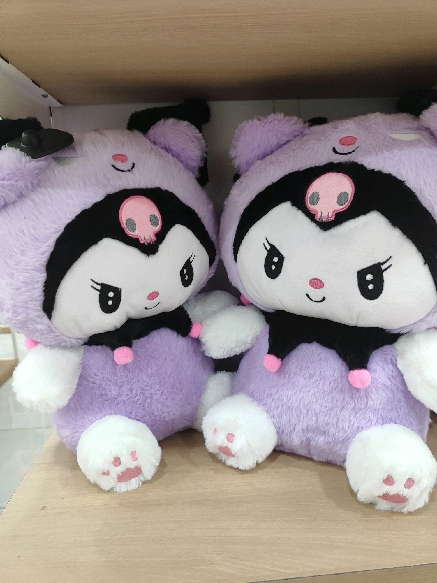 Two plush toys resembling the Sanrio character Kuromi are displayed side-by-side on a wooden shelf. The toys are predominantly lavender with black accents, featuring large white faces with black eyes, pink noses, and stitched mouths. Both have a pink skull motif on their foreheads, black ears, and white paws with pink paw prints. The left toy has a black ear tag. The background appears to be a light-colored wall or shelf. The image was taken in Sukowiryo, Indonesia.