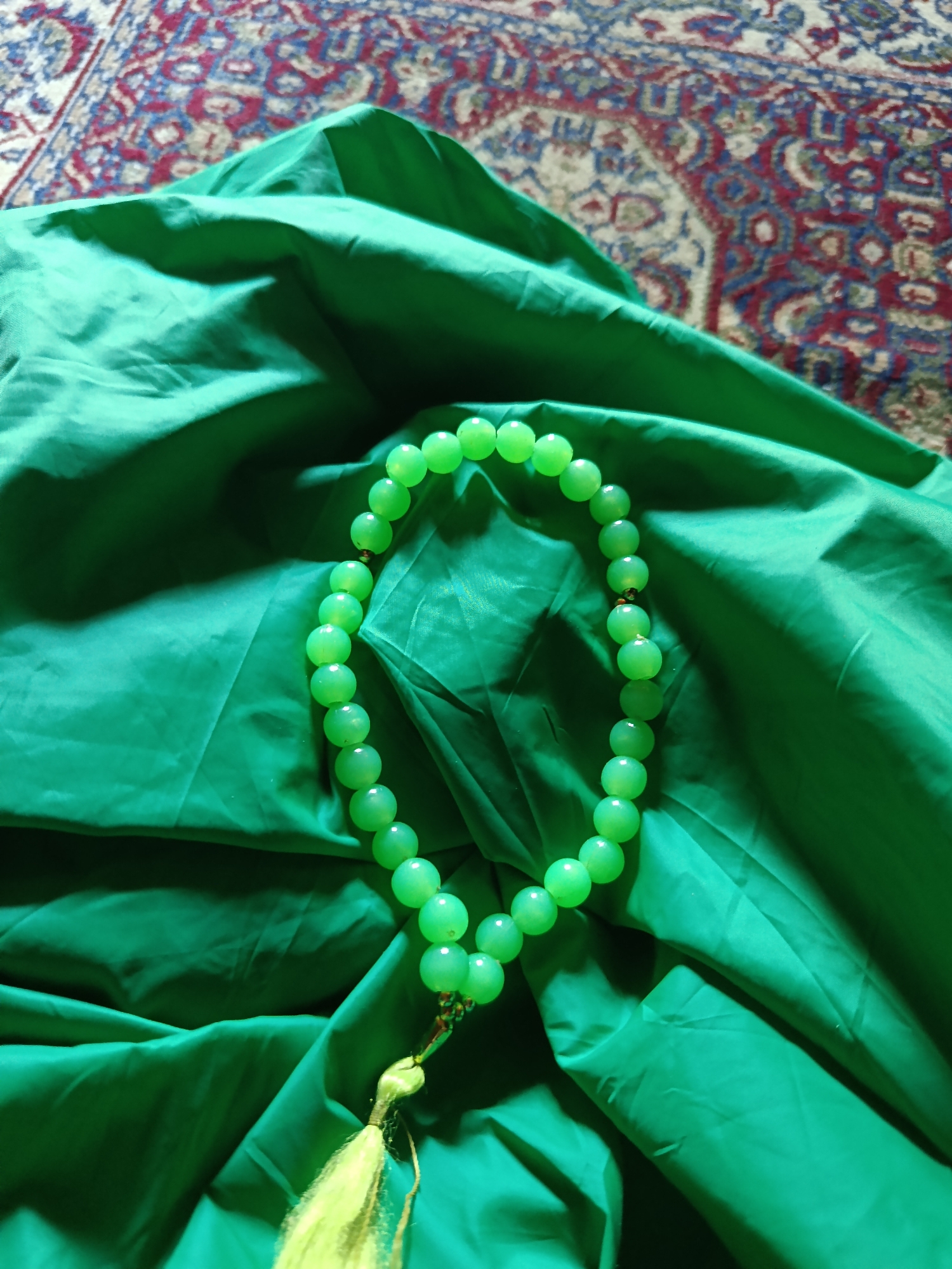 The image shows a close-up, overhead view of a string of light green prayer beads on a folded piece of emerald green fabric. The fabric is draped and has soft folds, suggesting it might be silk or satin. In the upper background, a portion of a rug with intricate red, blue, and cream-colored patterns is visible. The prayer beads are round and have a translucent quality, with a small golden bead separating sections of the green beads and a tasseled end with light yellow strings. The scene is lit from above, casting subtle shadows on the fabric. There are no people or discernible actions depicted. The location in Karangmojo, Indonesia, is not evident from the visual elements in the image.