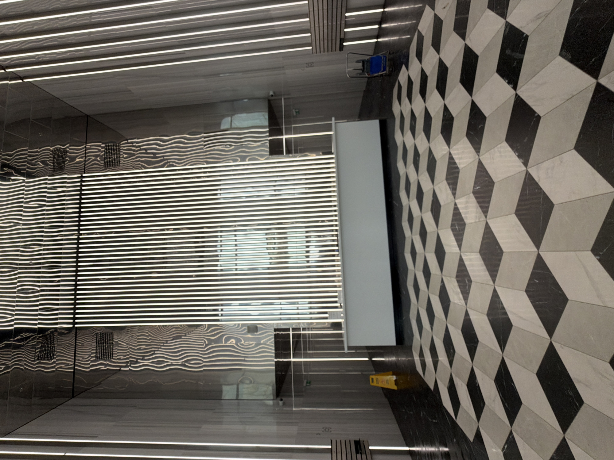 The image depicts the interior of a modern building lobby with a striking black and white geometric tiled floor. The floor tiles are arranged in a repeating pattern that creates an optical illusion of three-dimensional cubes or rhombuses, giving the impression of depth and an undulating surface.
Dominating the background is a wall featuring a series of vertical, illuminated elements that resemble fins or blinds, with a reflective surface behind them that distorts the light and creates wavy patterns. To the left and right, the walls are finished with dark, reflective material, likely marble or polished stone, interspersed with vertical LED strip lights.
In the mid-ground, a long, white counter or reception desk is positioned centrally. To the left of the counter, a yellow "wet floor" sign is visible on the dark part of the floor. In the distance, through doorways or archways, more of the building's interior can be seen, characterized by similar reflective surfaces and vertical lighting. A small blue cleaning cart is parked to the right of the counter.
There are no people visible in the scene. The overall impression is one of contemporary architectural design, emphasizing light, reflection, and geometric patterns. The image does not provide any specific clues to pinpoint the location to Moscow, Russia.