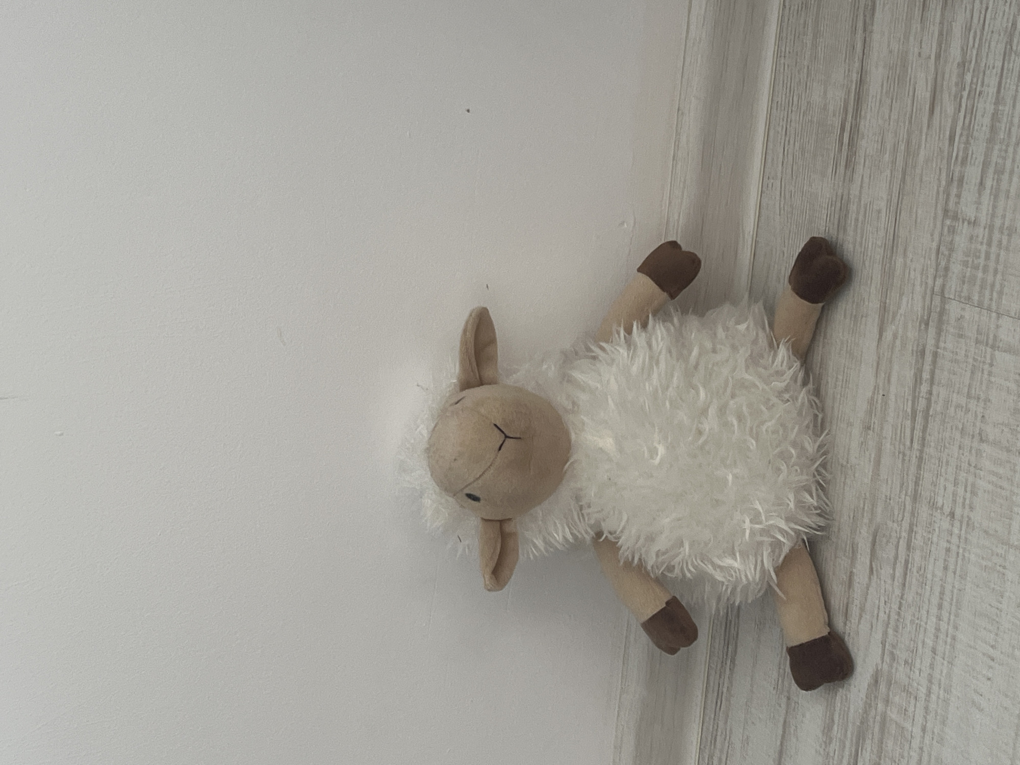 A plush lamb toy lies against a white wall and a section of light grey wood-patterned material. The toy has a round, beige head with small, floppy ears and simple stitched facial features. The body is covered in fluffy white material, and the legs are beige with brown feet. The toy is positioned so that its head is closest to the wall, and its legs extend towards the wood-patterned material. The scene suggests an indoor environment. It is not possible to determine if this scene is set in Blagoevgrad, Bulgaria from the given image.