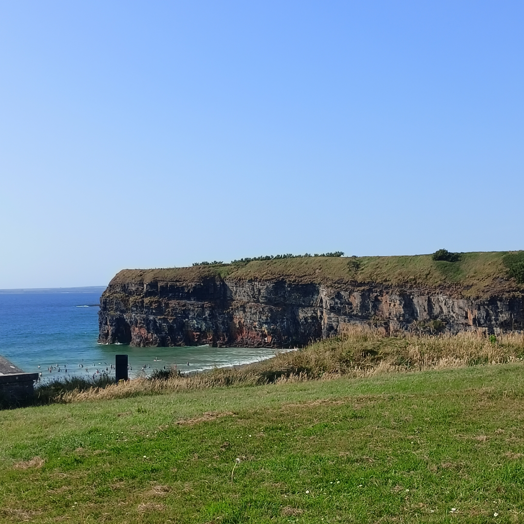 This is a photograph of the cliffs and beach at Ballybunion, Ireland, on a clear, sunny day. The foreground consists of a grassy hill sloping down towards a beach area. In the mid-ground, there are several people on the beach and in the water, some of whom appear to be surfing or swimming. The water is a vibrant turquoise color. Dominating the background are the impressive, layered cliffs, which are dark in color with patches of red and are covered in green vegetation at the top. The sky is a clear, light blue.