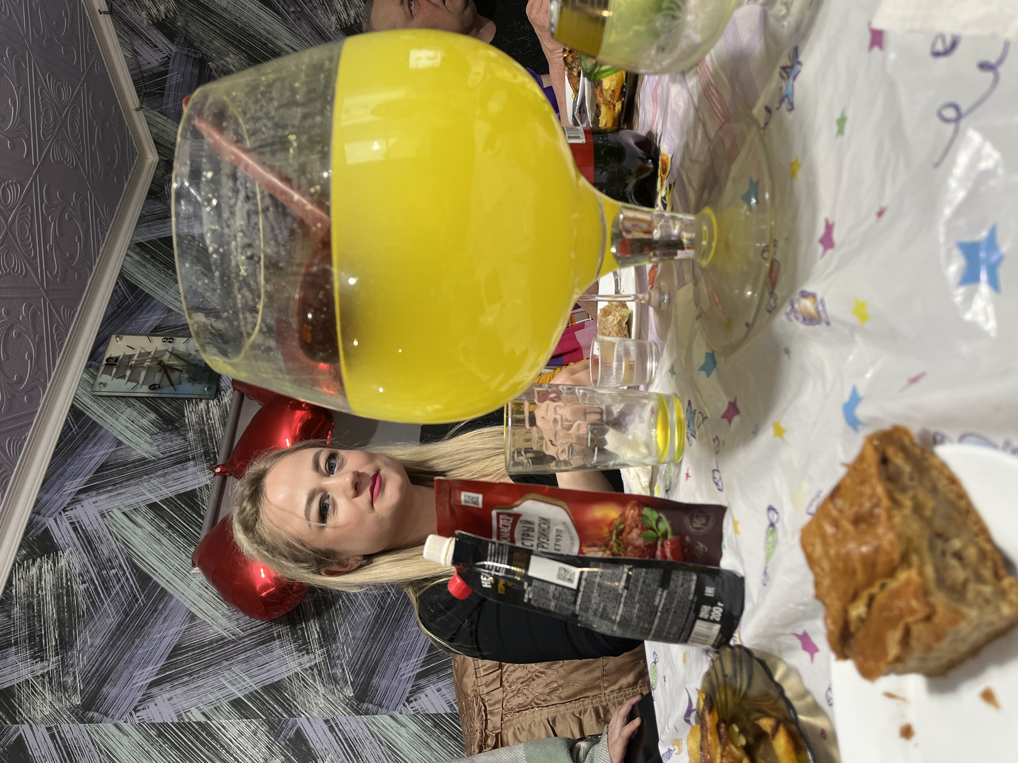 The image shows a woman with blonde hair sitting at a table. She is wearing a black jacket. On the table, there is a large glass of yellow liquid with a straw, a bottle of ketchup, a bottle of sauce, a plate with a piece of food, a small glass, and a white tablecloth with star patterns. In the background, there is a patterned wall. There are also red balloons visible. The location is likely Chaikovsky, Russia, due to the Cyrillic text on the sauce bottle. The image depicts a social gathering or meal.