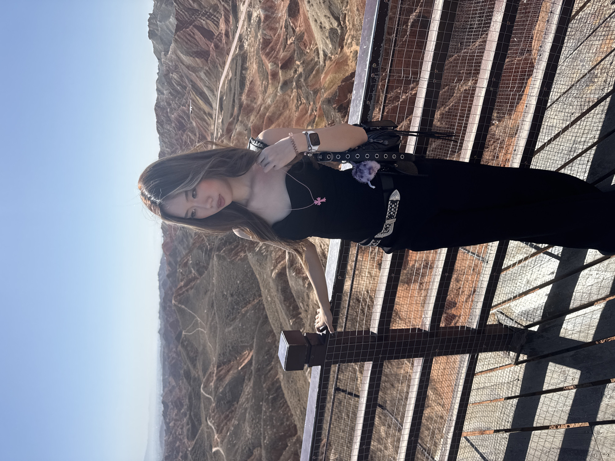 A young woman stands on an elevated outdoor walkway, posing with a vast, colorful mountain landscape in the background, under a clear, bright sky. The image is taken in direct daylight, indicated by the strong shadows cast by the walkway structure.
The woman, positioned slightly left of center, has long, dark hair with lighter highlights. She is wearing a black strapless top or dress, accessorized with a prominent belt featuring a large, ornate silver buckle. A light-colored watch is visible on her left wrist, and a small pink charm or pendant hangs from a necklace. A purple fluffy charm and a black bag with silver eyelets are also attached around her waist. Her expression is neutral as she looks towards the viewer. She is leaning with her right hand on a dark post that forms part of the walkway's railing.
The background reveals a spectacular geological formation, characterized by distinct layers of red, brown, and other earthy hues, indicative of Danxia landforms. These expansive, multi-textured mountains stretch across the horizon beneath a light blue sky. The walkway itself is constructed from metal grating and what appears to be wooden or metal planks, offering a panoramic view of the natural scenery. The overall scene suggests a visit to a scenic overlook in a mountainous region, specifically Zhangye, China, known for its unique Danxia landscapes. There is no visible text.