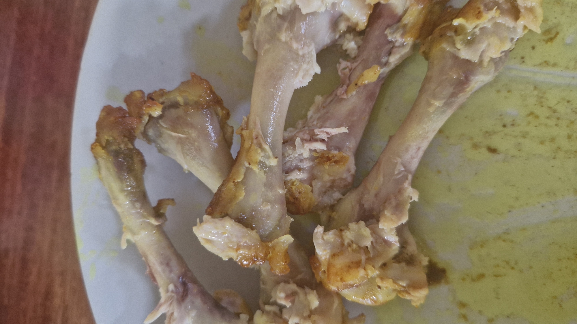 The image shows a close-up view of several cooked chicken bones and scraps on a white plate with a light yellow residue. The bones are light brown and have some remaining meat attached. The plate is on a wooden surface, likely a table. The lighting is overhead, casting some shadows. The scene is static, with no visible movement or activity. The location is not specified beyond "Unknown, Unknown."