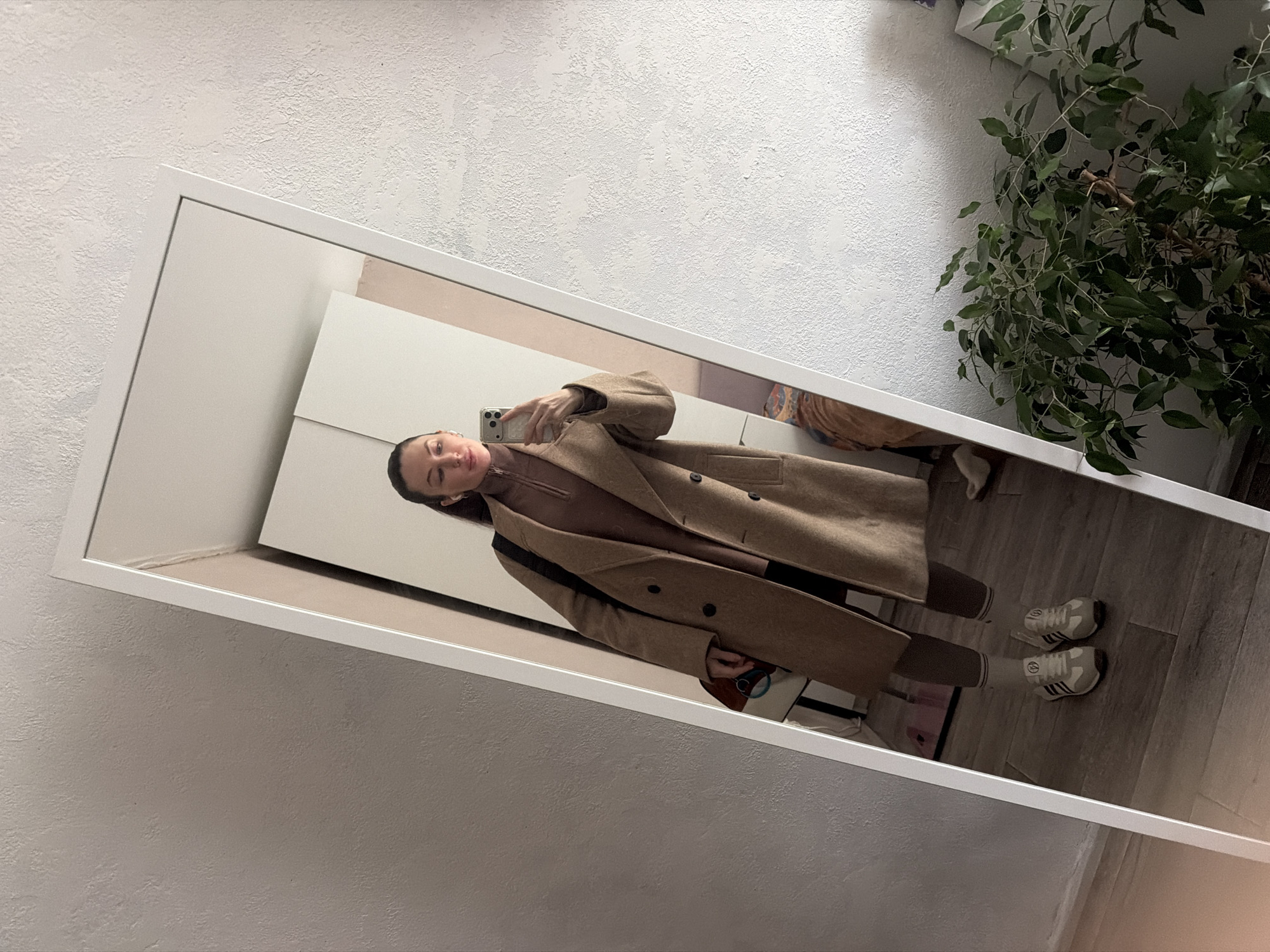 A woman is reflected in a tall rectangular mirror. She is holding a smartphone in her right hand, presumably taking a picture of herself. She is wearing a brown turtleneck and a long, double-breasted beige coat with dark buttons. She also wears dark leggings and beige sneakers with stripes around the ankles.
The mirror is leaning against a white wall with a textured surface. In the background, partially visible, is what appears to be a white cabinet. A potted plant is on the floor near the mirror. The floor has a wood-like surface. The location is unspecified and is not definitively identified as the city of Perm, Russia.
