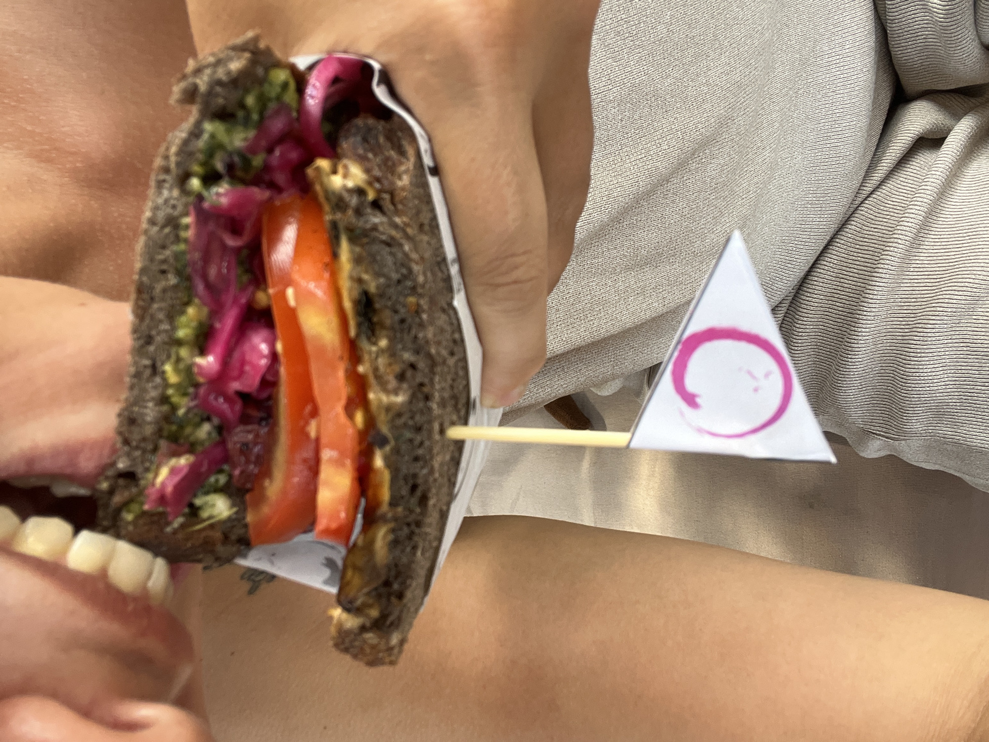 A person is about to bite into a sandwich. The sandwich is held in one hand and has dark bread, green spread, purple onions, and red tomatoes. A white triangular flag with a pink circle is attached to the sandwich with a toothpick. The person's mouth is open, revealing teeth. The person is wearing beige clothing. The location is the Ubud District, Indonesia.