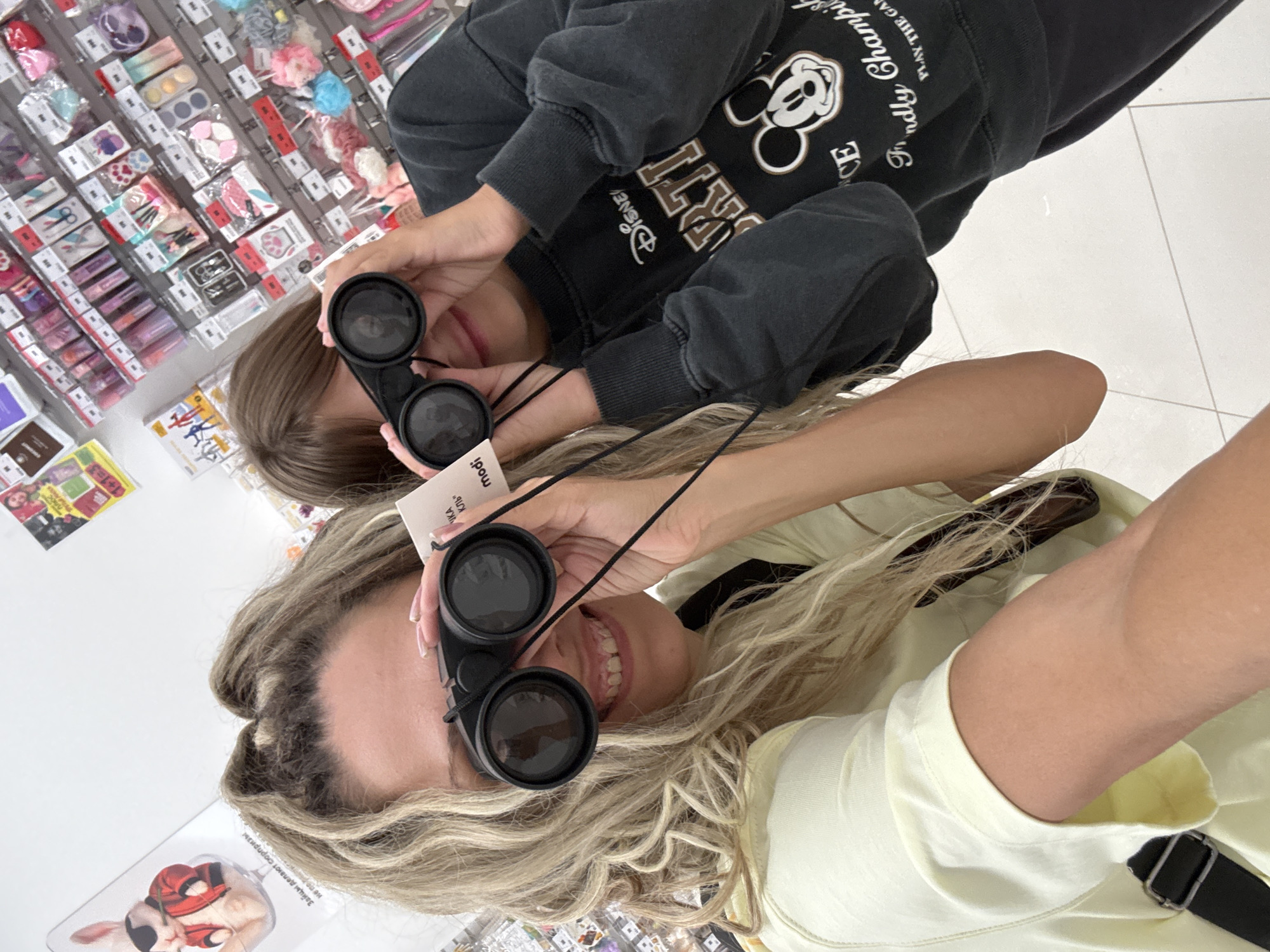 Two individuals, one adult and one child, are posing for a selfie in a retail store. The adult, with long blonde hair, is in the foreground, wearing a light yellow t-shirt. They are holding a black pair of binoculars up to their eyes and smiling. The child, in the background, is wearing a dark grey sweatshirt with a Disney Mickey Mouse graphic and is also holding binoculars to their eyes. The store appears to be a novelty or gift shop, with shelves stocked with various items like plush toys, hair accessories, and other small trinkets. The lighting is bright and artificial, consistent with an indoor retail environment. The floor is tiled. There is no visible indication that this location is in Sosnovtsy, Russia.