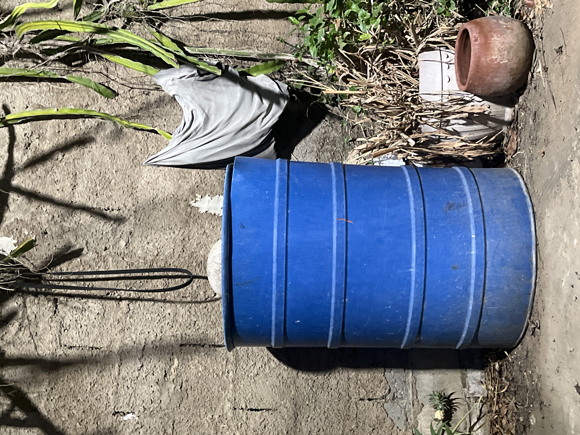 A blue plastic barrel stands in front of a textured concrete wall. To the right of the barrel are several potted plants, one containing dried vegetation and another a small reddish-brown pot. Above the barrel, hanging from the wall, is a black strap and a piece of grey fabric draped over a vine. The lighting suggests it is nighttime, with shadows cast on the wall from overhead elements. The ground appears to be a mixture of dirt and gravel. The scene is located outdoors, in an urban environment, likely a yard or alleyway, in Santa Ana, Venezuela.