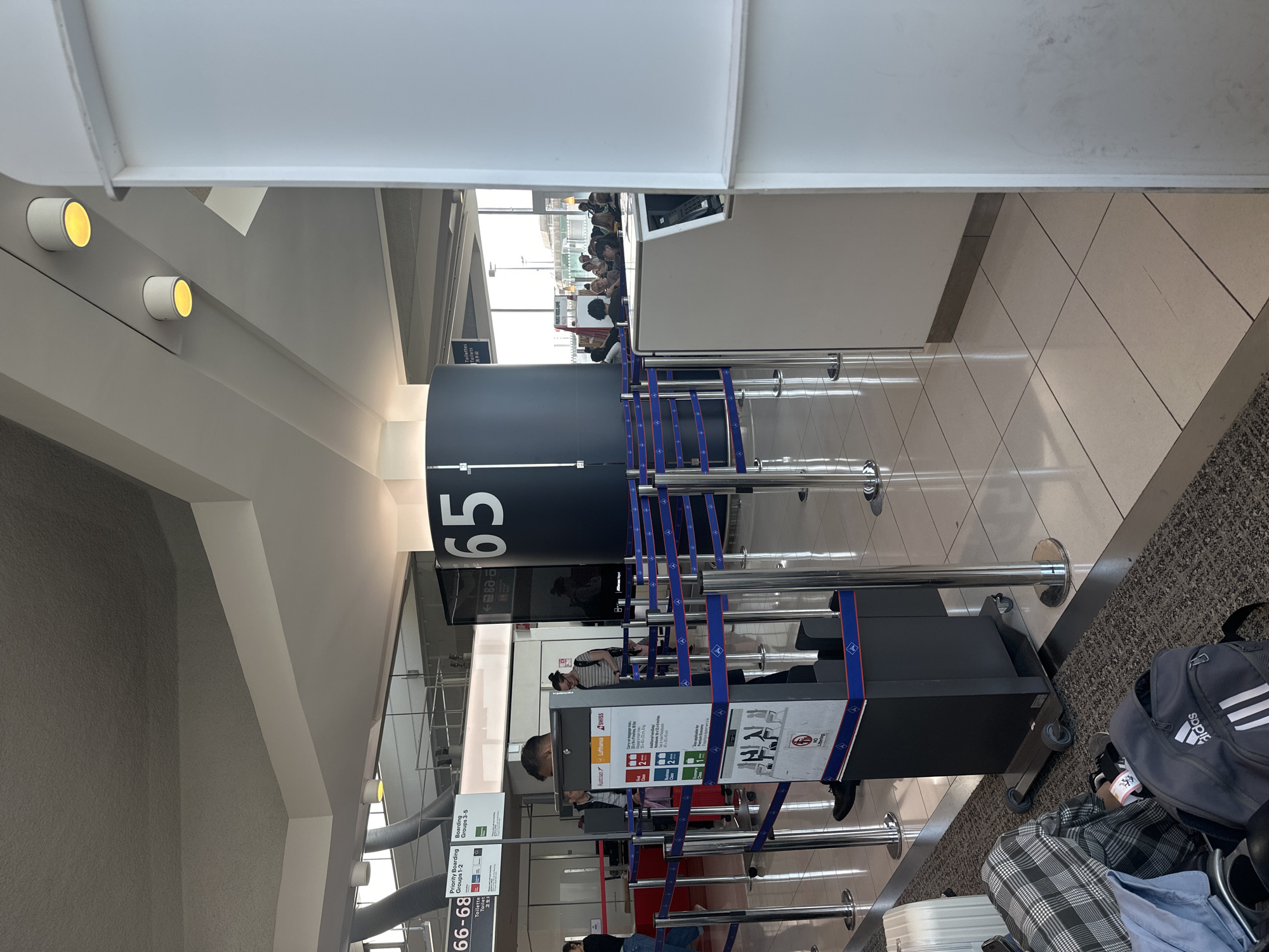 The image captures an airport boarding gate area, specifically Gate 65, in Mauregard, France. The scene shows multiple travelers either queuing or waiting near the gate.
In the center, a large, dark cylindrical sign prominently displays the gate number "65" in white lettering. Blue and white stanchions create defined queues, leading towards self-service boarding kiosks. Several people are visible: some are standing in line, one person appears to be interacting with a kiosk, and others are seated further back. Luggage, including a grey Adidas backpack, a plaid carry-on bag, and a white suitcase, rests on the tiled floor and a carpeted area.
Visible text includes "65" on the main gate sign, and smaller signs providing boarding information such as "Priority Boarding Groups 1-2" and "Boarding Groups 3-5". Airline logos and names for "Austrian", "Lufthansa", and "SWISS" are also displayed on the kiosks, indicating flights for these carriers. Additional signage points to "Toilette" or "Toilets" and a gate range "66-68" in the background.
The setting features modern airport architecture with white walls, a structured ceiling, and recessed warm yellow lights. The flooring consists of light-colored tiles and a grey carpet. The atmosphere appears calm, typical of passengers waiting for their flight.