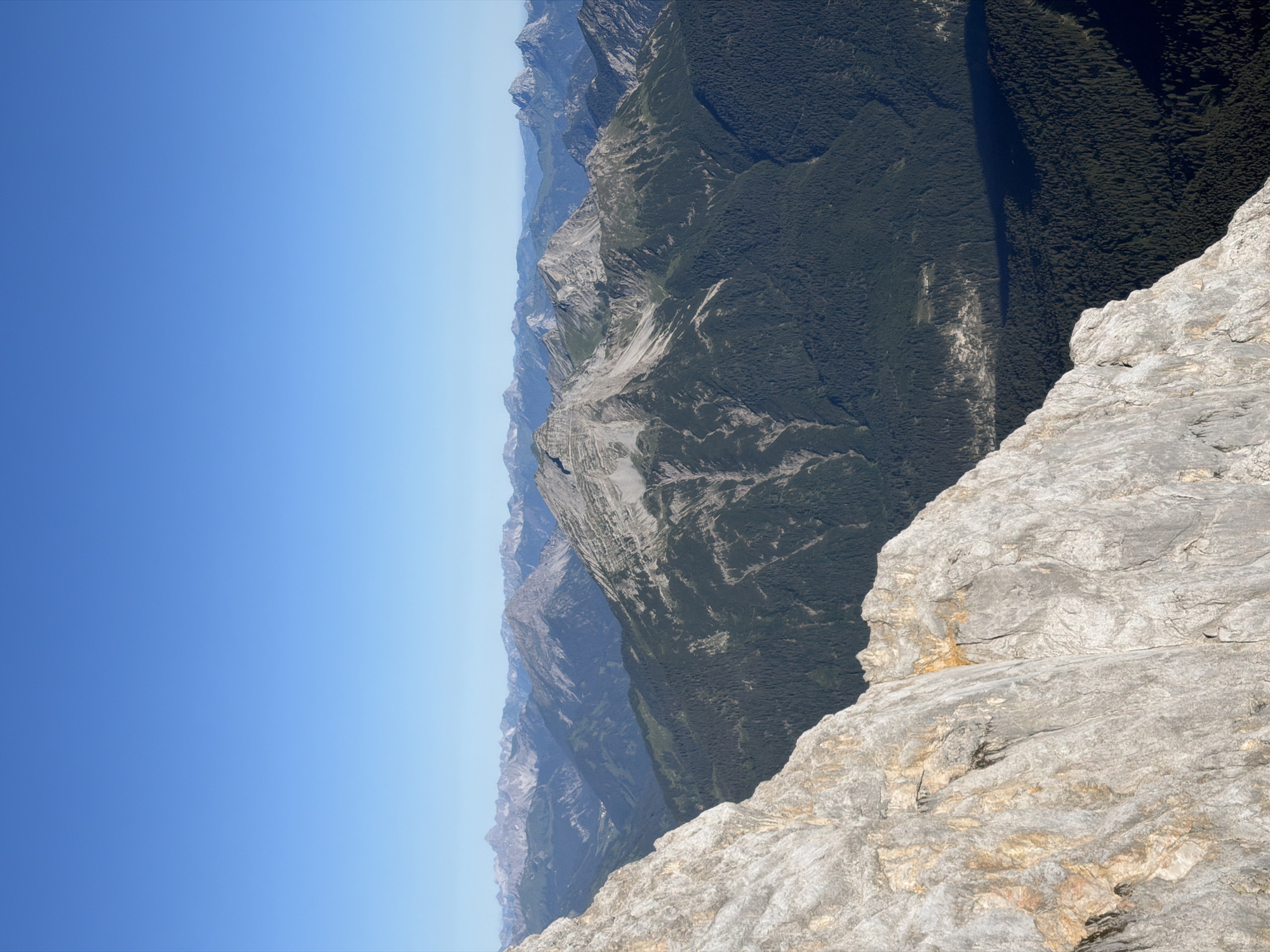The image is a panoramic view of a mountainous landscape with a clear blue sky. In the foreground, a large, rocky cliff face dominates the left side of the frame, showing texture and geological stratification with patches of yellow and orange discoloration. Beyond this cliff, a vast expanse of rugged mountains stretches into the distance. These mountains are characterized by steep slopes covered in dense, dark green coniferous forests, interspersed with lighter green alpine meadows and rocky peaks. The distant mountain ranges fade into a hazy blue, suggesting a significant distance and clear atmospheric conditions. The lighting suggests it is daytime, likely morning or afternoon, casting shadows across the valleys and highlighting the topography of the terrain. There are no people or man-made structures clearly visible in the immediate foreground or mid-ground, giving the scene a sense of natural wilderness. The location is likely Grainau, Germany, given its proximity to the Bavarian Alps, known for such dramatic mountain scenery.