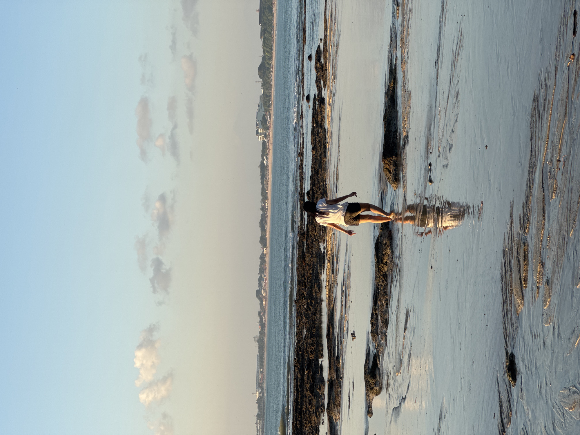 A young woman walks on a wet sandy beach in Kuta Selatan, Indonesia, during the golden hour. The sky is a soft blue with scattered white and gray clouds. The ocean is calm and blue, with a gentle wave breaking on the left side of the image. The beach is covered in dark, wet sand and large, dark rocks. The woman is facing away from the camera, wearing a white t-shirt and dark shorts. She is reflected in the shallow water on the sand. In the background, a long, sandy coastline with buildings and trees is visible across the water.