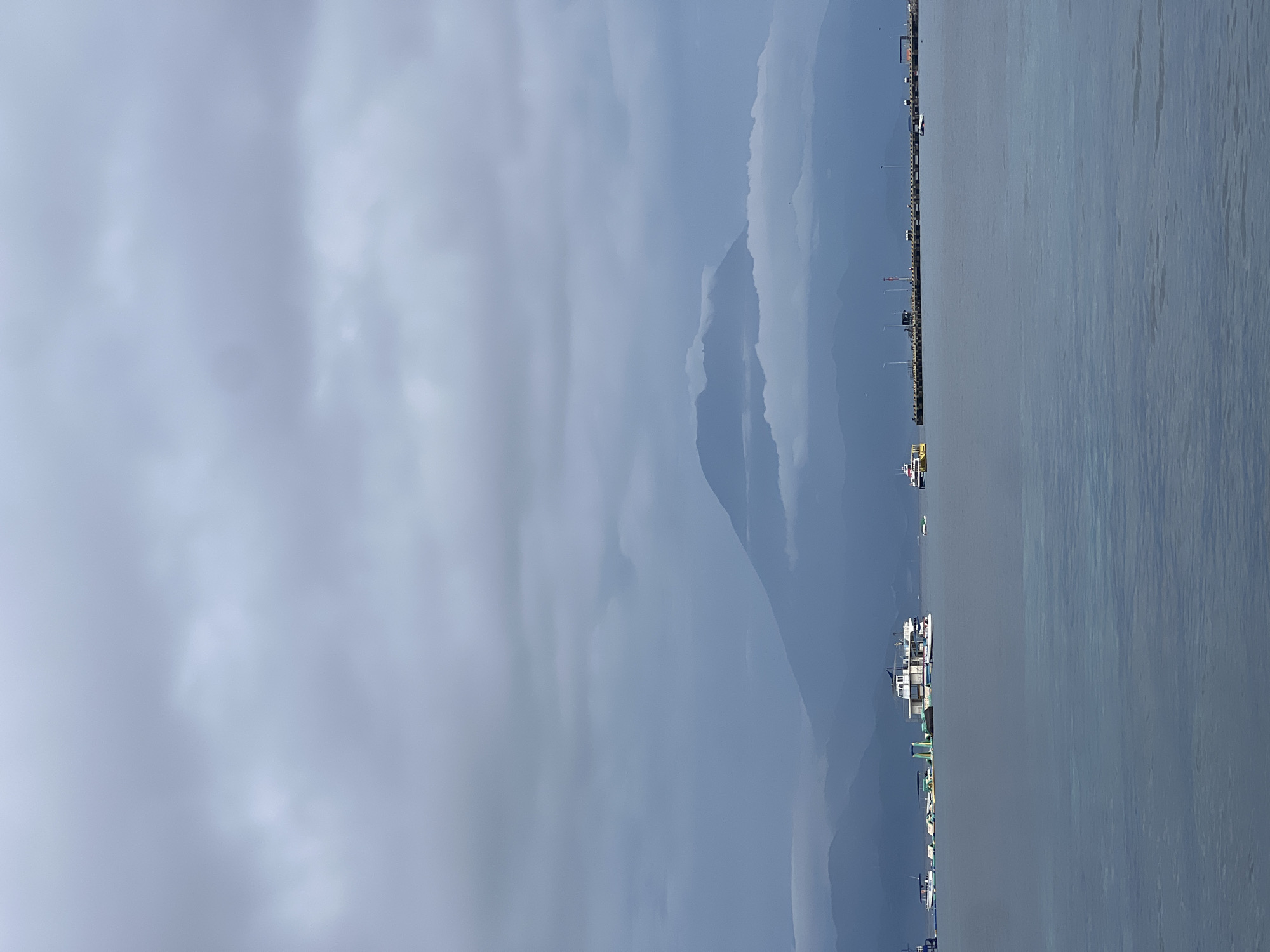 The image presents a tranquil coastal landscape under an overcast sky. A wide expanse of calm, grey-blue water dominates the right half of the frame, showing very subtle ripples on its surface. The left half is filled with a soft, diffused grey sky, heavily laden with clouds that suggest an overcast or hazy day, providing a uniform, subdued light to the entire scene.
Along the horizon, which horizontally divides the sky and water, lies a long, narrow strip of land that appears to be a pier or a coastal embankment. This structure is lined with indistinct small buildings or shelters, poles, and what appear to be lights, stretching into the distance. In the far background, behind this coastal strip and beneath the cloud-covered sky, multiple layers of mountains or large hills are visible. Their silhouettes are softened by atmospheric haze, appearing as progressively lighter shades of blue-grey as they recede.
Several boats are scattered across the water. A notable white and blue boat with a multi-level cabin is visible in the middle-left section of the water, positioned near the pier. Further along the pier, towards the background, another smaller, lighter-colored boat can be seen. Additional faint specks in the far distance on the water might indicate other distant vessels.
The scene captures a peaceful moment in a maritime setting, likely off the coast of Nusa Penida, Indonesia, as suggested by the location context. The overcast conditions create a serene and slightly muted atmosphere. No discernible text is visible within the image.