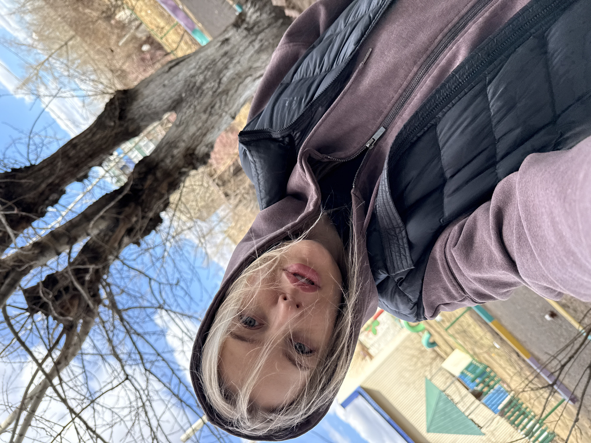 This close-up, angled selfie features a young person, likely female, with light blonde hair, blue eyes, and light skin, looking directly into the camera with a neutral expression. She is wearing a dark purple or mauve hooded sweatshirt, with the hood pulled up around her face, over a black quilted puffer vest or jacket. The outerwear is partially unzipped.
The background depicts an outdoor scene under a clear blue sky with some wispy clouds. Large trees with bare, dark branches and rough trunks are prominent on the left side of the frame, suggesting a season of late autumn, winter, or early spring. To the right and bottom, various ground elements are visible, including what appears to be part of a colorful outdoor mat or paved area with abstract shapes and outlines, hinting at a playground or recreational space. Faint structures or buildings can be seen in the distant background.
The image is significantly rotated, giving a skewed perspective where the sky is in the upper left corner and the ground slopes towards the lower right. The natural light indicates it is daytime, and the layered clothing suggests cool weather. No visible text is present in the image. The location is Borzya, Russia, and the activity is the individual taking a self-portrait outdoors.