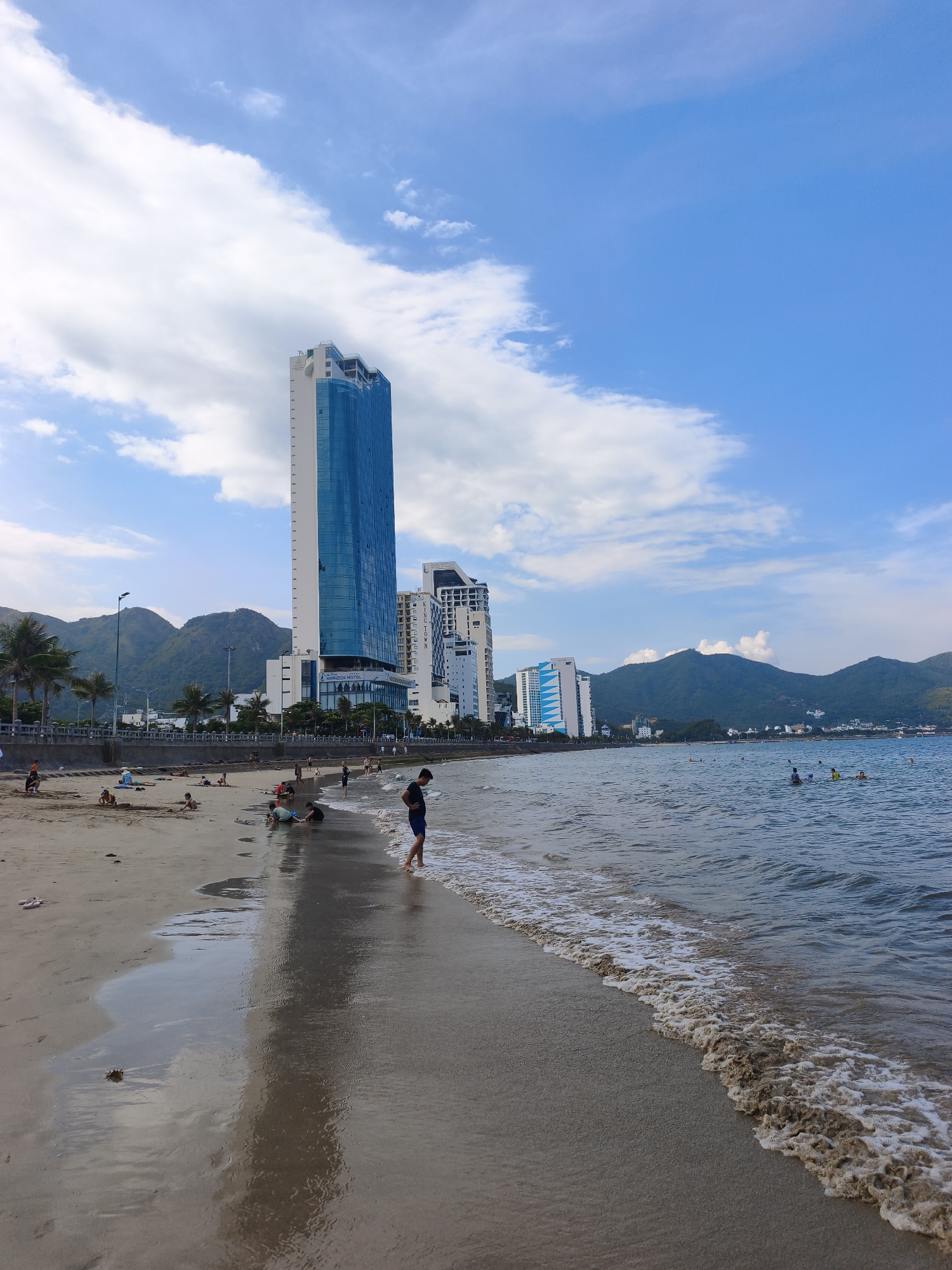 This image depicts a scenic beachside view in Nha Trang, Vietnam, likely along Tran Phu Beach. In the foreground, a sandy beach meets the ocean with gentle waves lapping at the shore. A man stands near the water's edge on the right, facing away from the camera. Further up the beach, several people are scattered, some engaging in activities like building sandcastles, while others relax. Palm trees line the walkway behind the beach, leading to a line of modern high-rise buildings. Dominating the cityscape is a tall, slender skyscraper with a blue glass facade, its height suggesting a prominent hotel or residential tower. Behind the buildings and palm trees, lush green mountains form a dramatic backdrop under a clear blue sky dotted with fluffy white clouds. The scene captures a typical day at this popular coastal destination, blending natural beauty with urban development.
