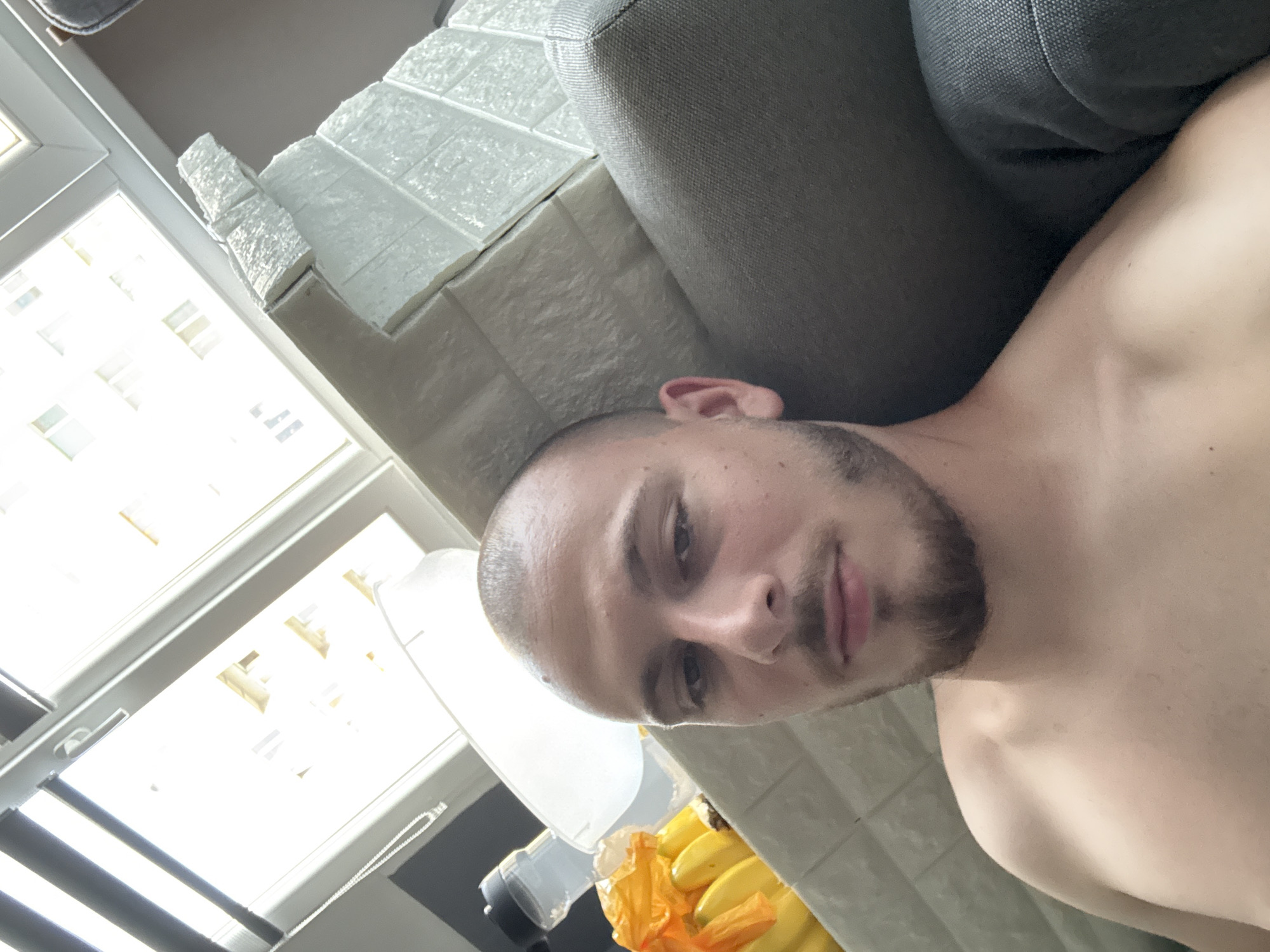 The image is a selfie taken by a young man with a shaved head and a short beard. He is shirtless and leaning against a gray sofa. To his left, there is a collection of bananas in an orange plastic bag and a water bottle. Behind him, there is a window with natural light streaming in, illuminating a wall with textured white panels resembling bricks. The background suggests an indoor setting, possibly an apartment in Stara Zagora, Bulgaria, given the architectural style visible through the window. The overall lighting is bright due to the natural light source.
