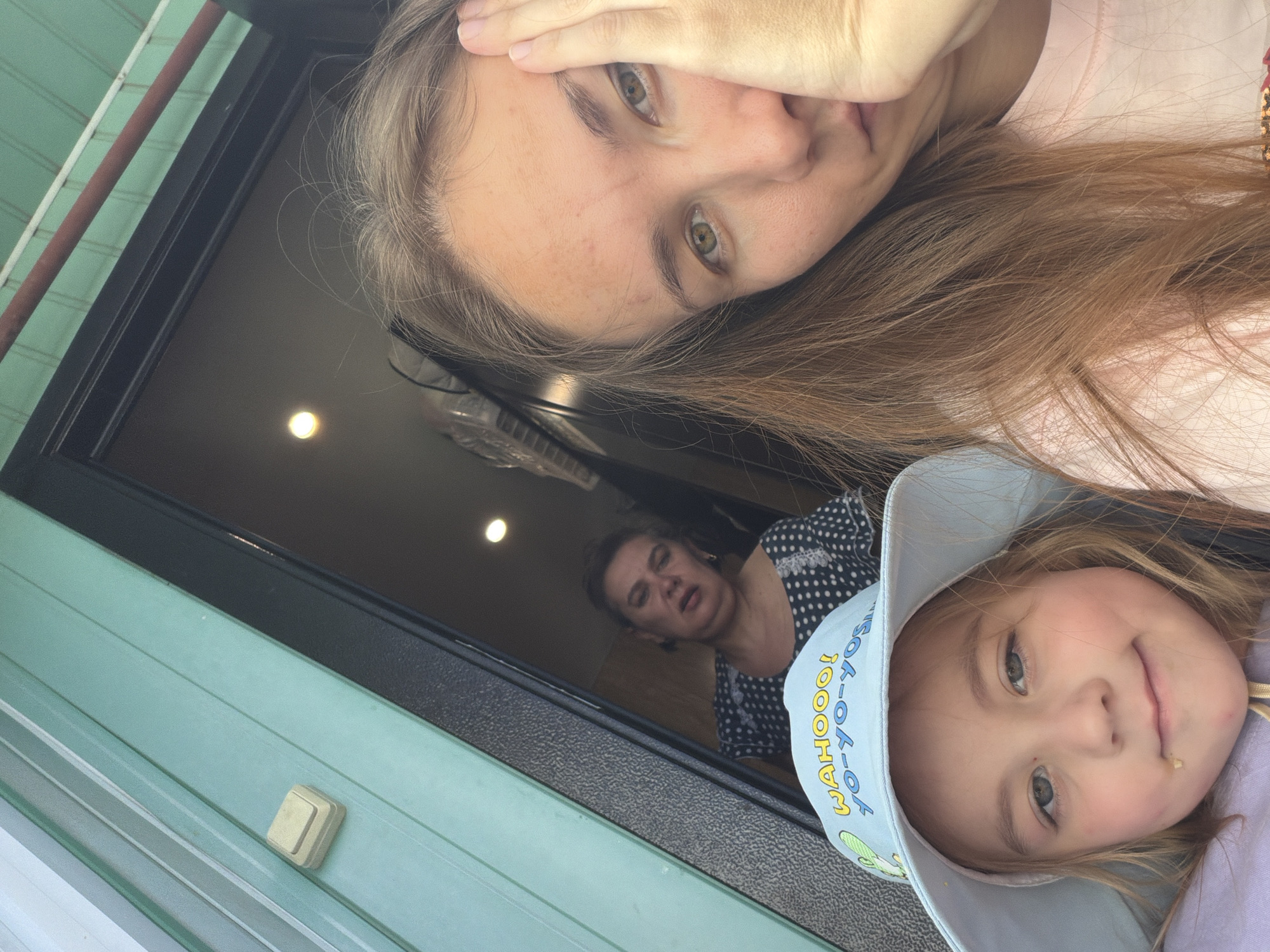 In Tambov, Russia, an outdoor selfie captures three females in close proximity to an entryway. On the left, a young girl wearing a light blue bucket hat with "WAHOOO! YO-YO-YO-YO-YO!" and cartoon imagery is prominently featured. To her right, a woman is captured from the chest up, her face partially obscured by her hand; she appears to be looking towards the camera with a pensive expression. Behind them, a third woman, positioned further back in the entryway, is visible, her face angled away from the camera, appearing to be speaking or reacting to something off-camera. The environment suggests a dwelling, with a teal-colored wall on the left and a dark doorway opening into a dimly lit interior. The overall scene is casual and personal.