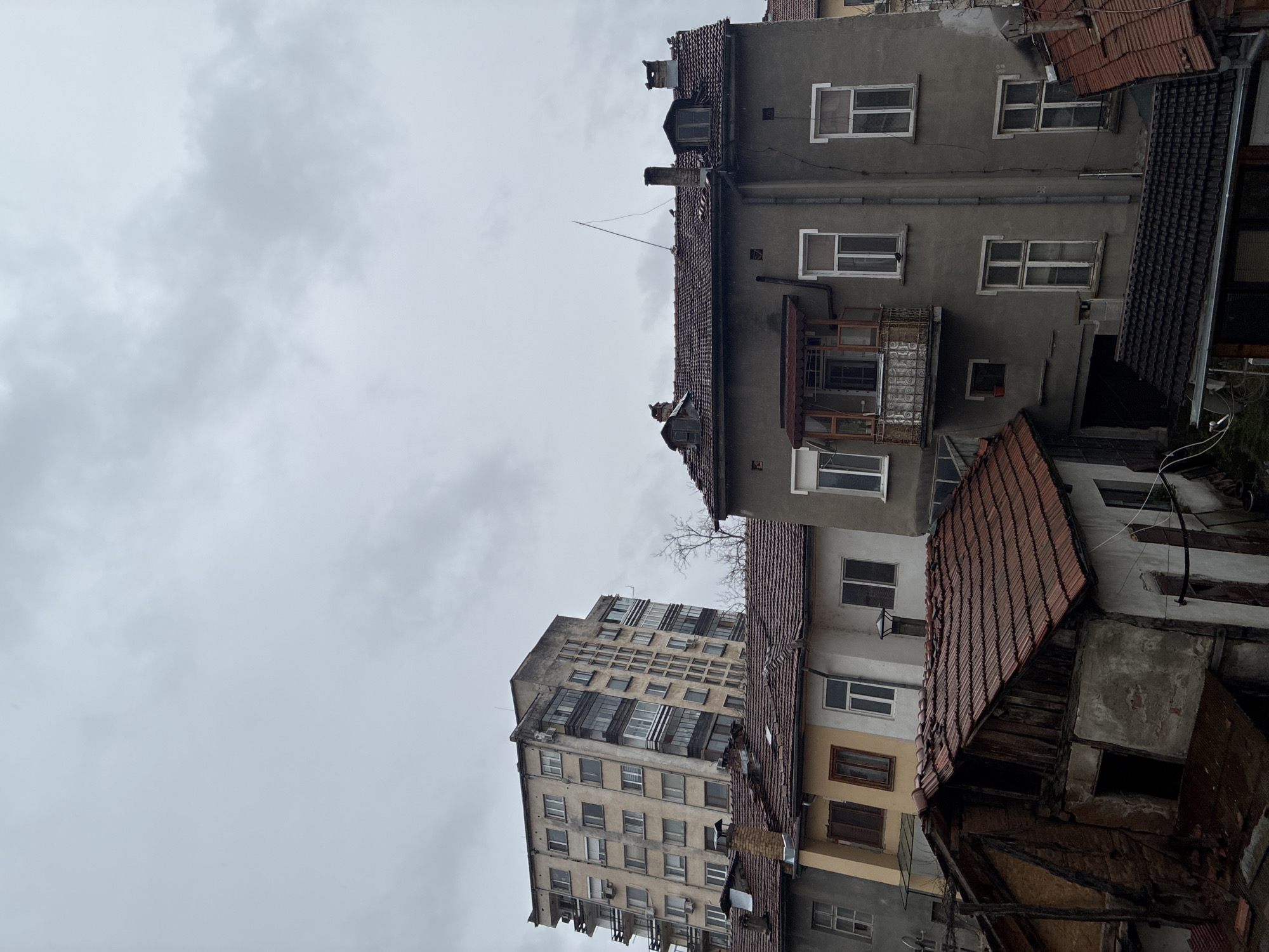 The image depicts a view of buildings in Troyan, Bulgaria, under an overcast sky. Dominating the scene are several aging buildings of varying heights and architectural styles. The building closest to the viewer is multi-storied, with multiple windows. The roofs are tiled. The overall environment suggests an older, possibly historic, part of the city. No people are visible in the frame.